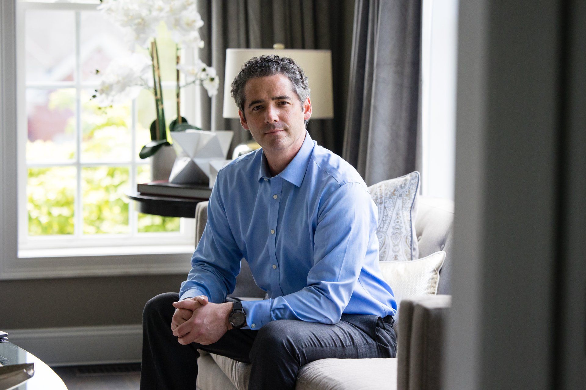Man sitting on a couch in a bright living room near a window and white flowers