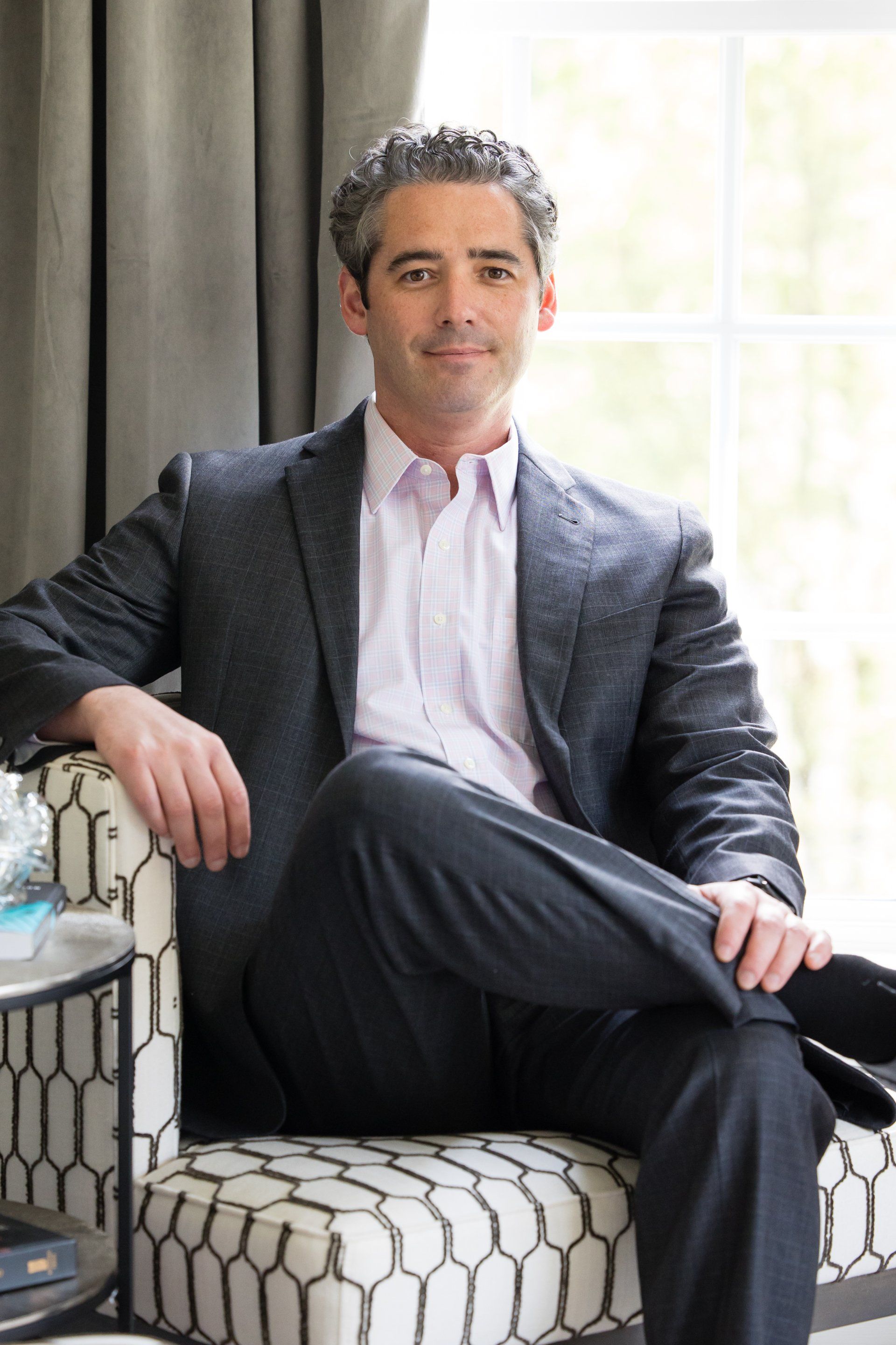 Man seated in a patterned chair by a window, wearing a dark blazer and light shirt, legs crossed.