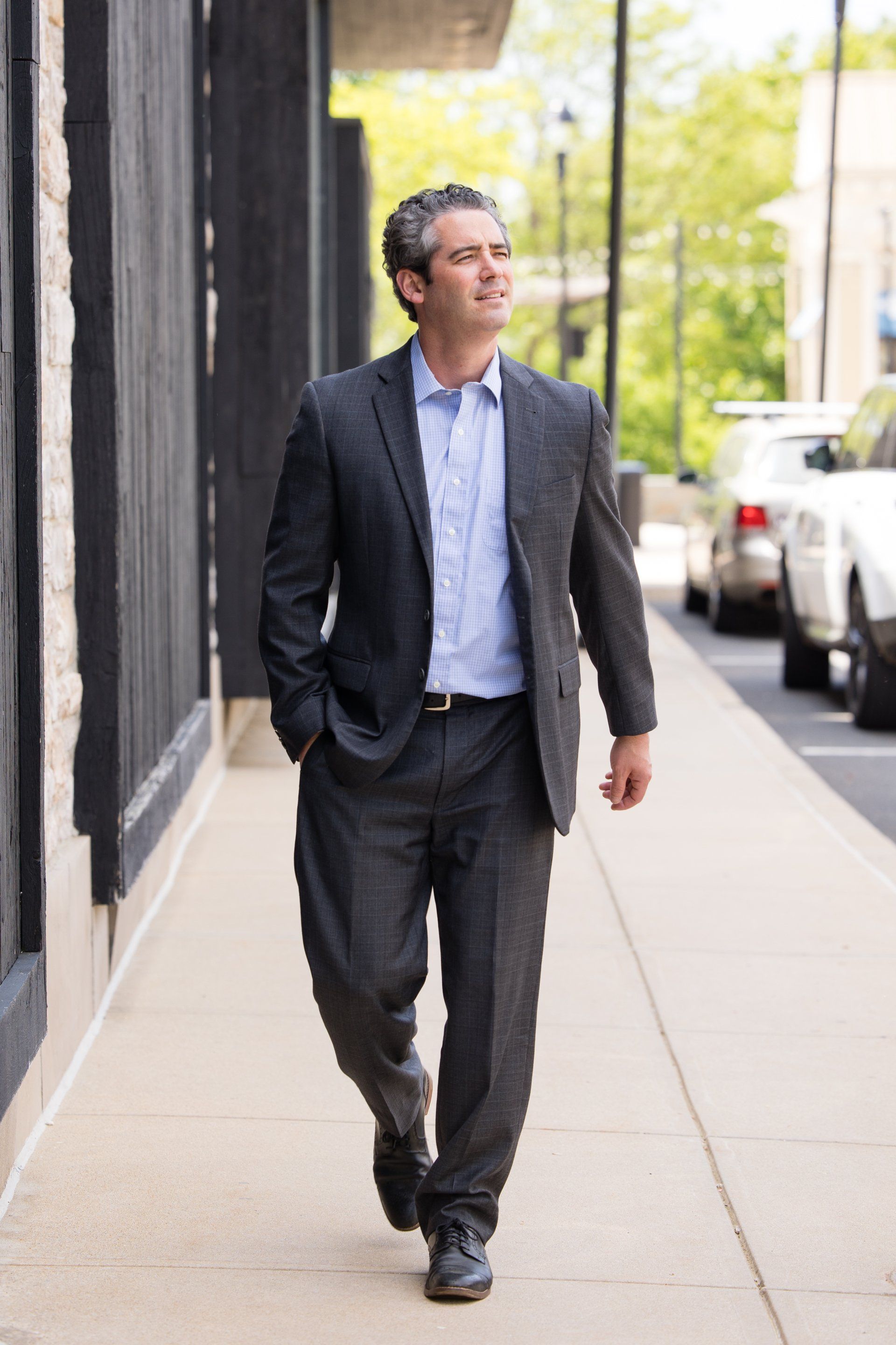 Man in a dark suit walking along a city sidewalk beside parked cars and buildings.