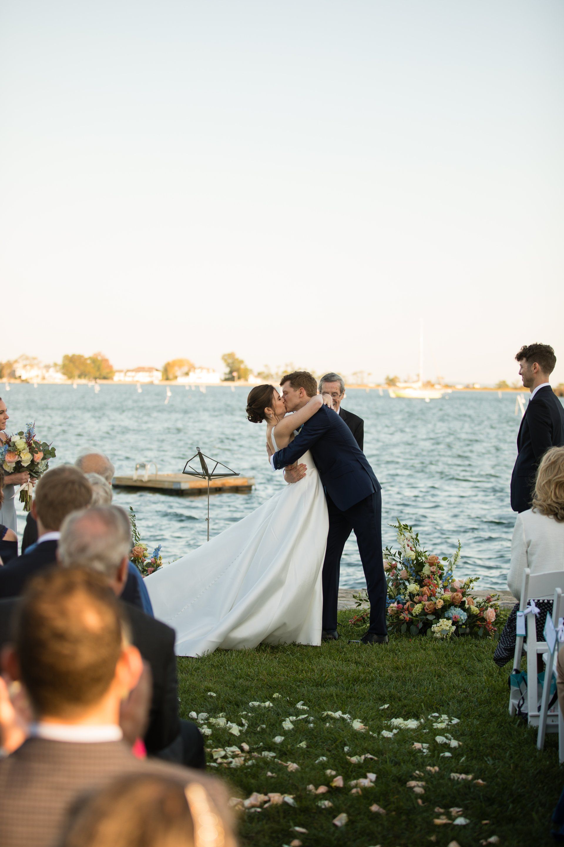 Wedding couple kissing by the water at sunset, with guests seated nearby.