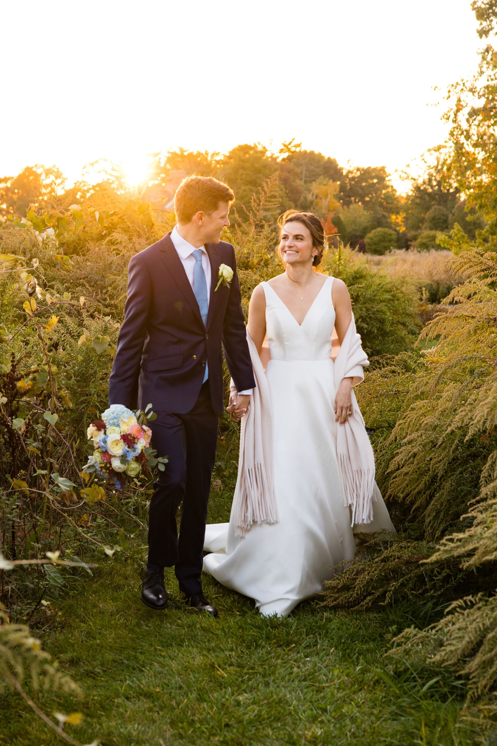 Newlyweds walking in a sunlit garden, holding hands and a bouquet, with warm evening light.