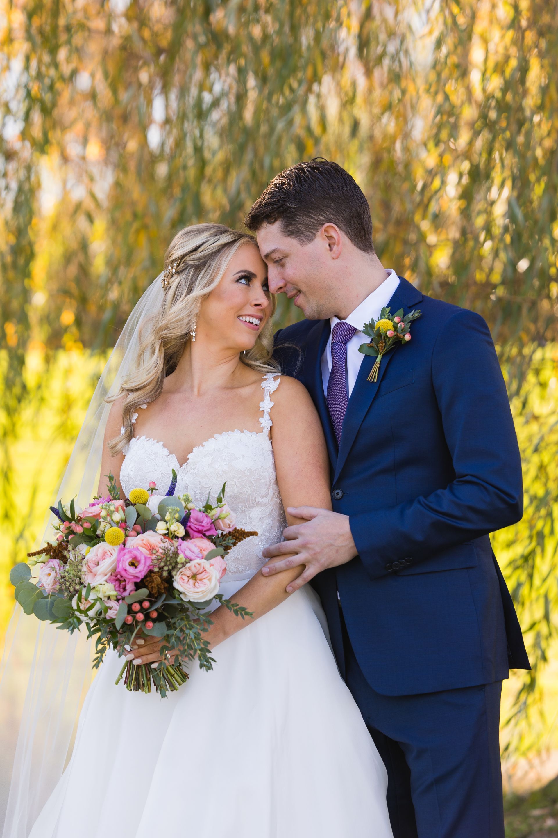 Bride and groom in blue suit and white gown holding a bouquet, standing outdoors by willow trees