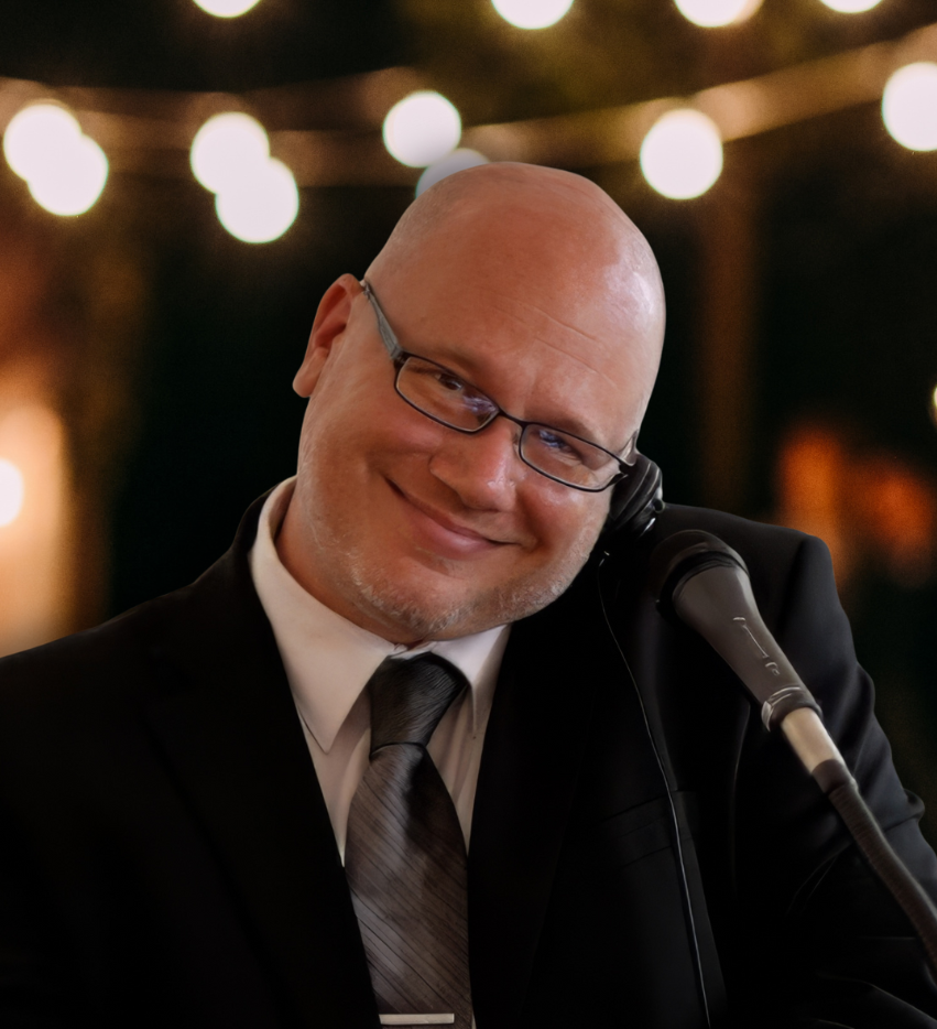 Smiling bald man in glasses beside a microphone, wearing a suit and tie at a warmly lit event
