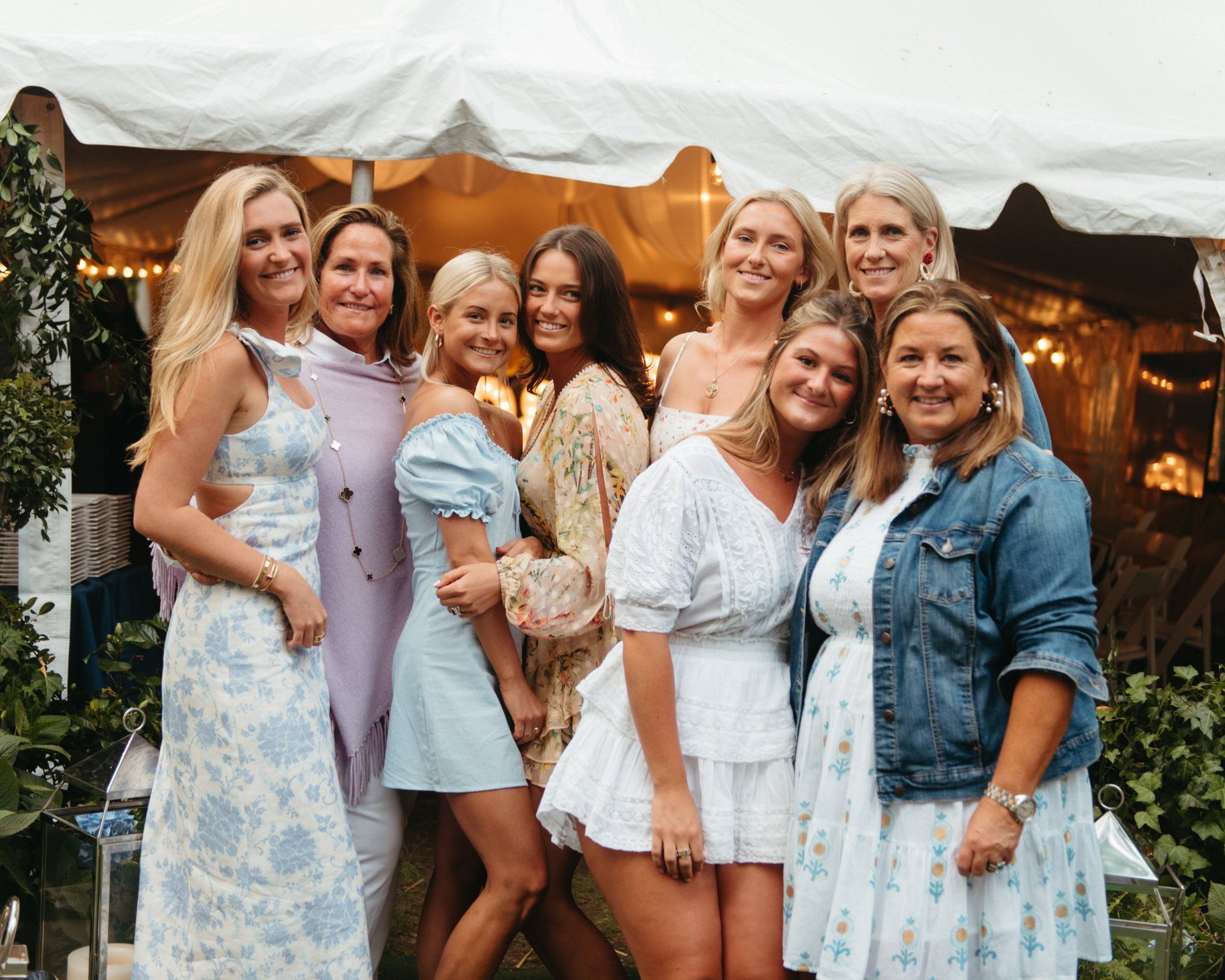 Seven women posing together at an outdoor event in light blue and white dresses, smiling under a tent