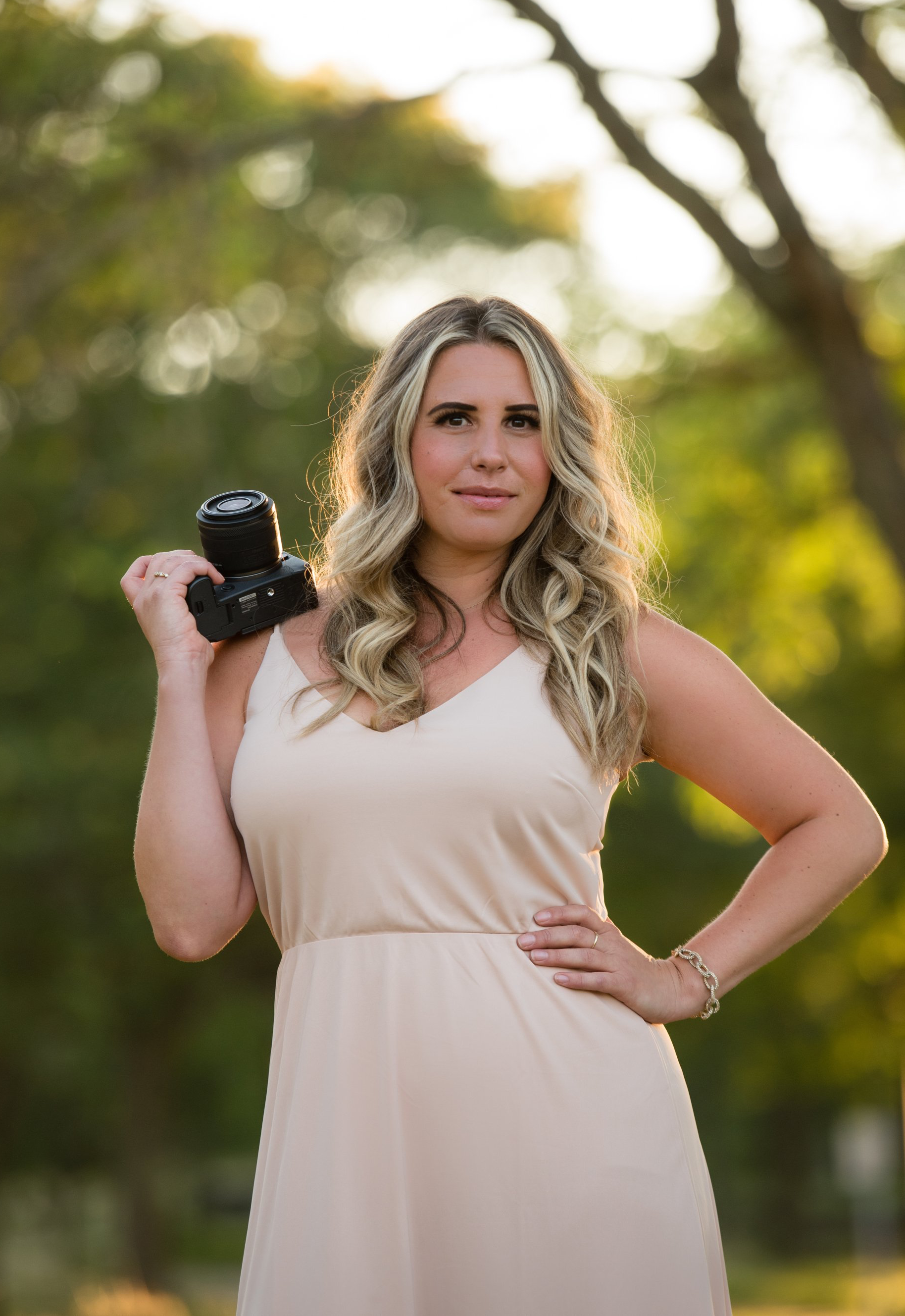 Woman in a beige dress holding a camera outdoors with a blurred green background