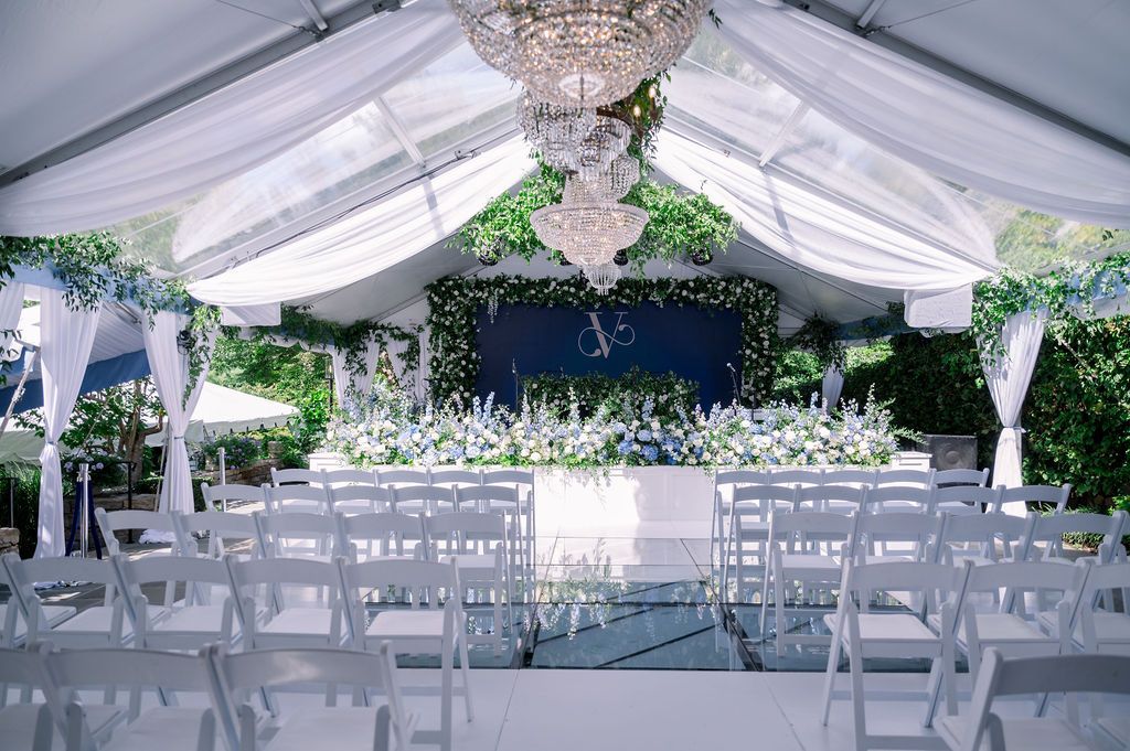 White tented wedding reception with chandeliers, floral decor, and rows of white chairs facing a stage