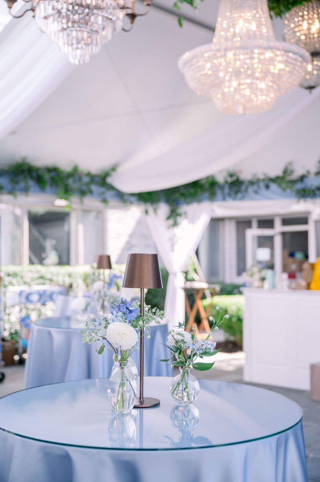 Elegant outdoor reception table with blue glass top, floral centerpiece, and chandeliers under a white tent
