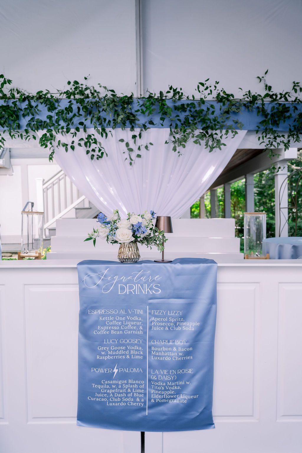 Blue wedding seating chart with white text, greenery, and white draped decor at an indoor reception table