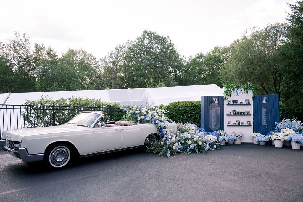 Vintage white convertible decorated with blue-and-white flowers beside a draped outdoor event backdrop