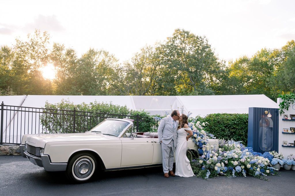 Classic white convertible at an outdoor wedding with a bride, groom, and blue-white floral arch