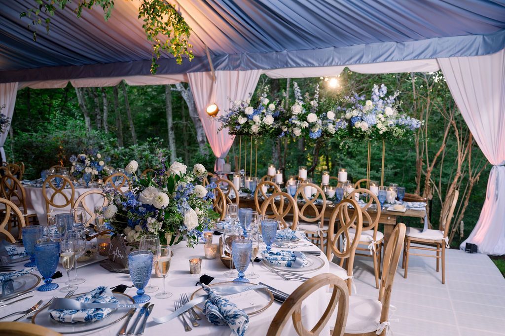 Elegant outdoor banquet under a draped tent with blue-and-white table settings, floral decor, and warm string lights