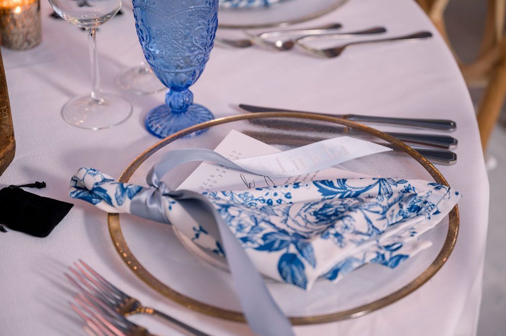 Elegant table setting with white plate, blue floral napkin, gold rim, and silverware on a white tablecloth