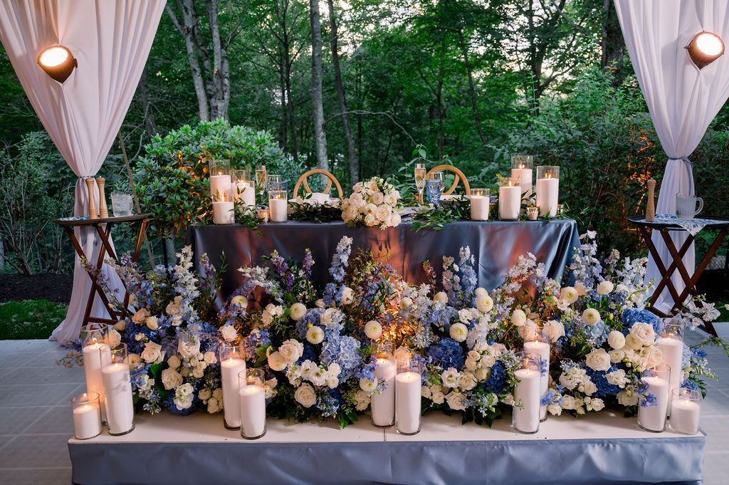 Outdoor floral wedding altar with candles, white drapes, and lush blue-white flower arrangements in a garden setting