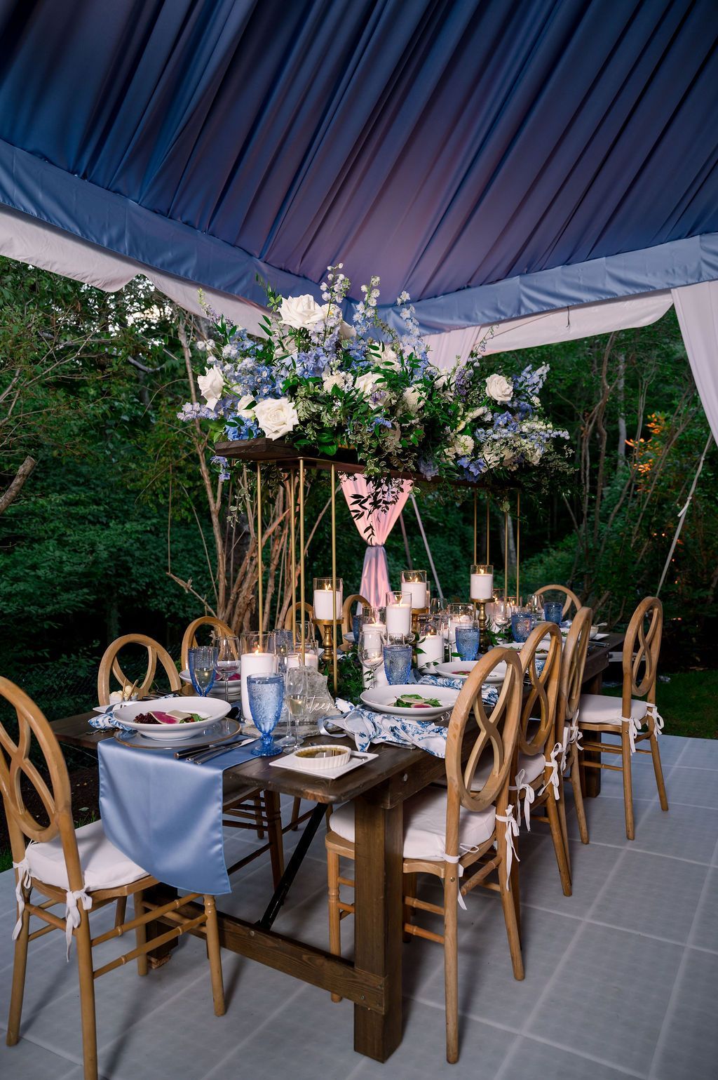 Elegant outdoor dining table under a tent, set with flowers, candles, and wooden chairs at dusk
