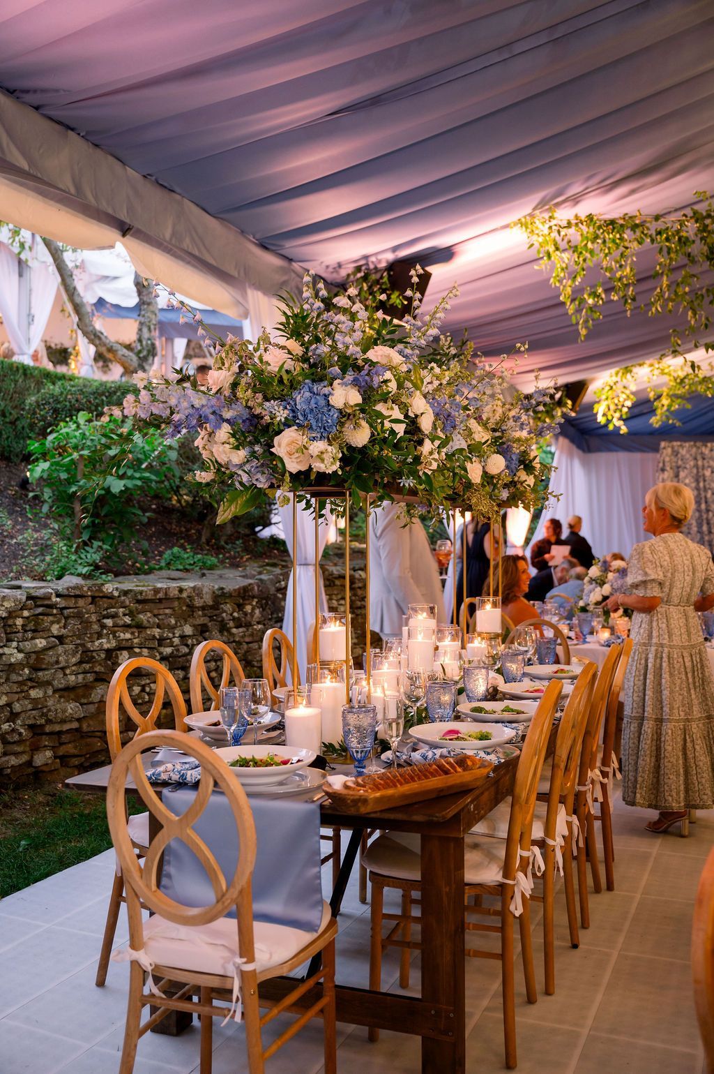 Elegant outdoor banquet table under a tent with white chairs, candles, and floral arrangements
