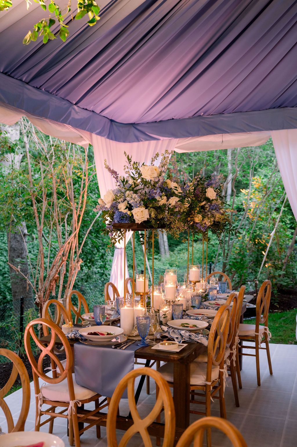 Outdoor banquet table under a lavender tent with floral centerpiece and candles, set in a garden
