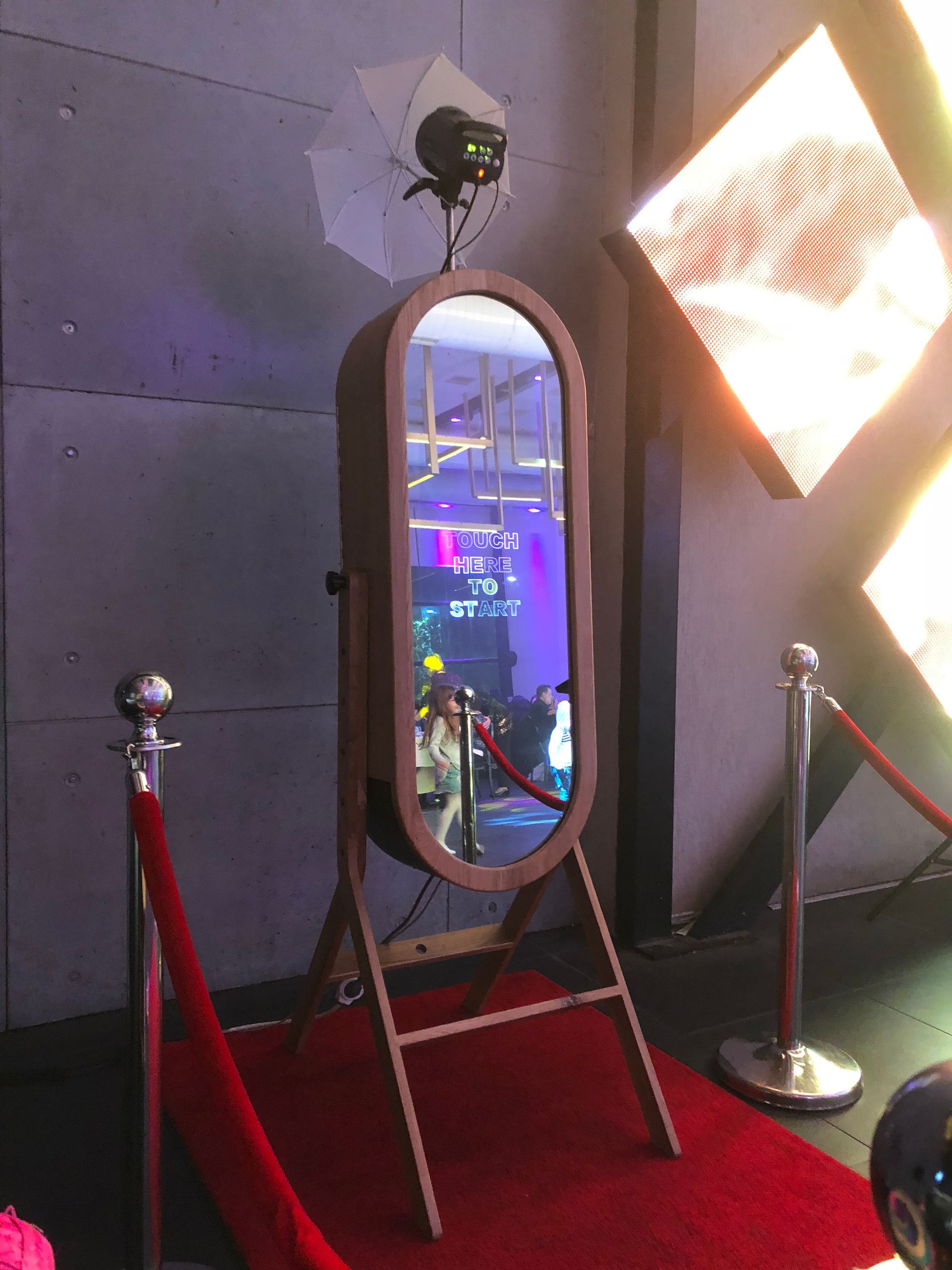 Tall oval mirror on a stand in a dim lounge with red carpet and velvet ropes, reflecting neon lights.