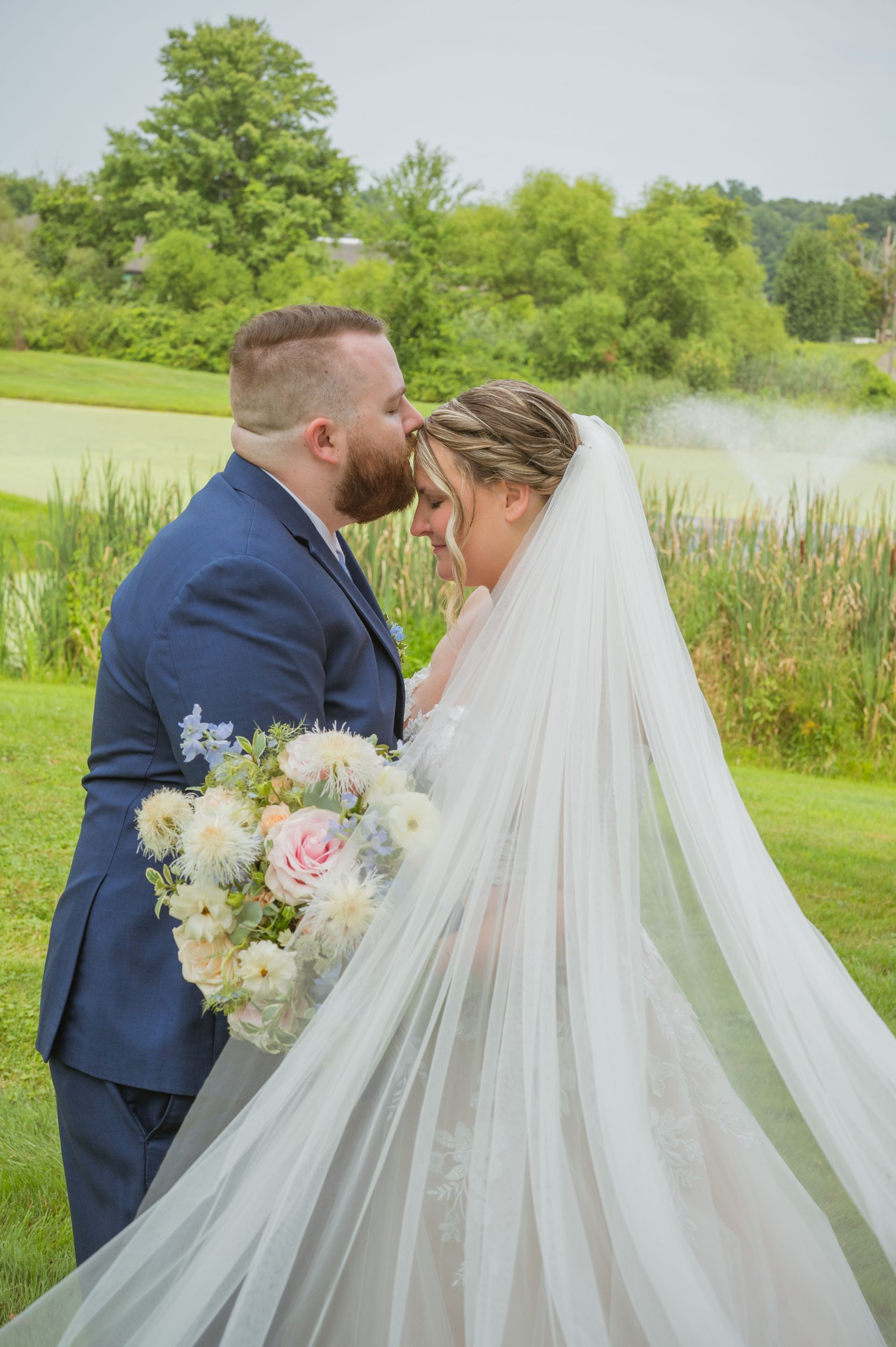 Bride and groom kissing by a lake, with the bride’s long veil flowing and a bouquet in hand