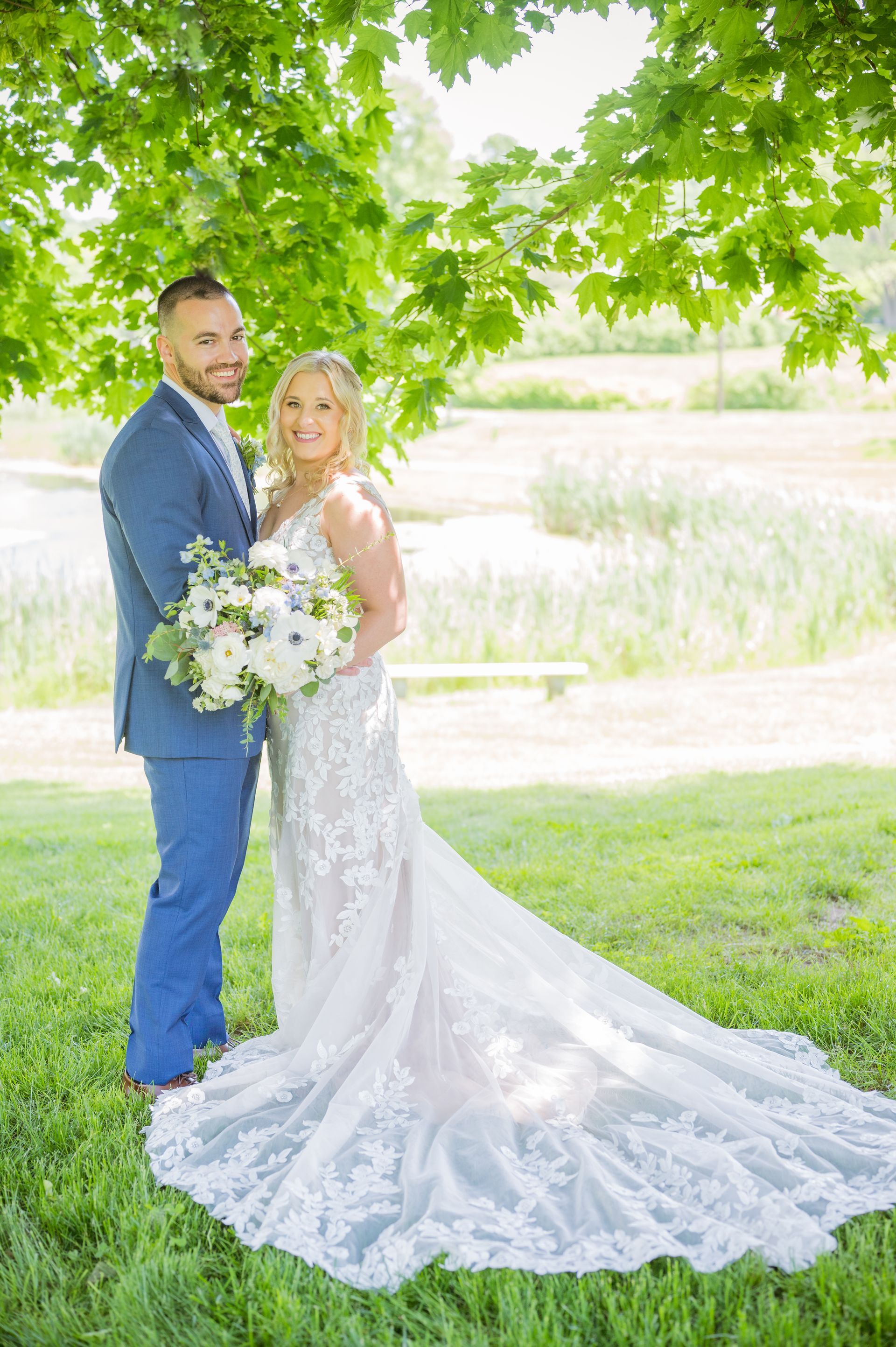 Bride and groom standing under leafy trees, the bride in a white gown holding a bouquet outdoors