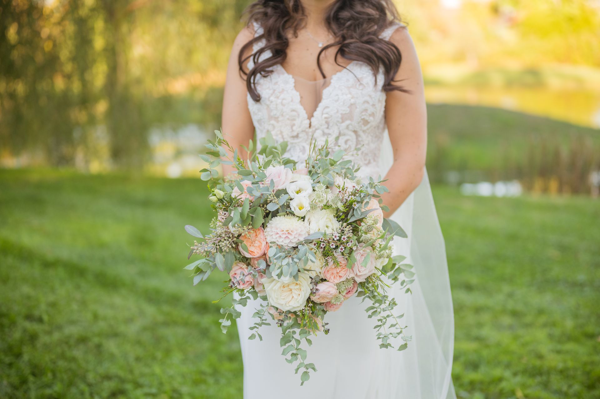 Bride holding a pastel bouquet in a white lace wedding dress outdoors on a green lawn