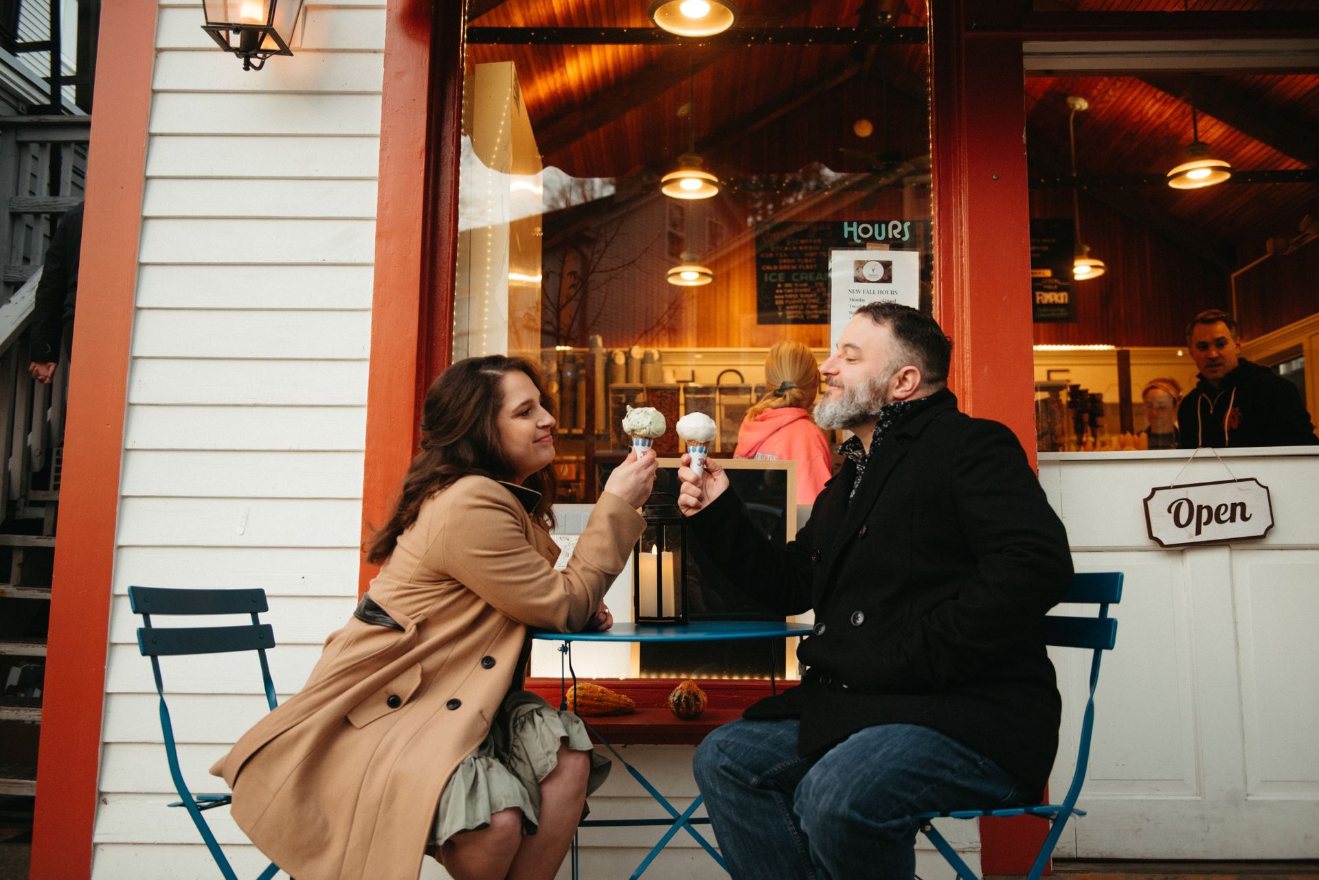 Two people clink wine glasses at a café patio, seated by a storefront with warm lights.