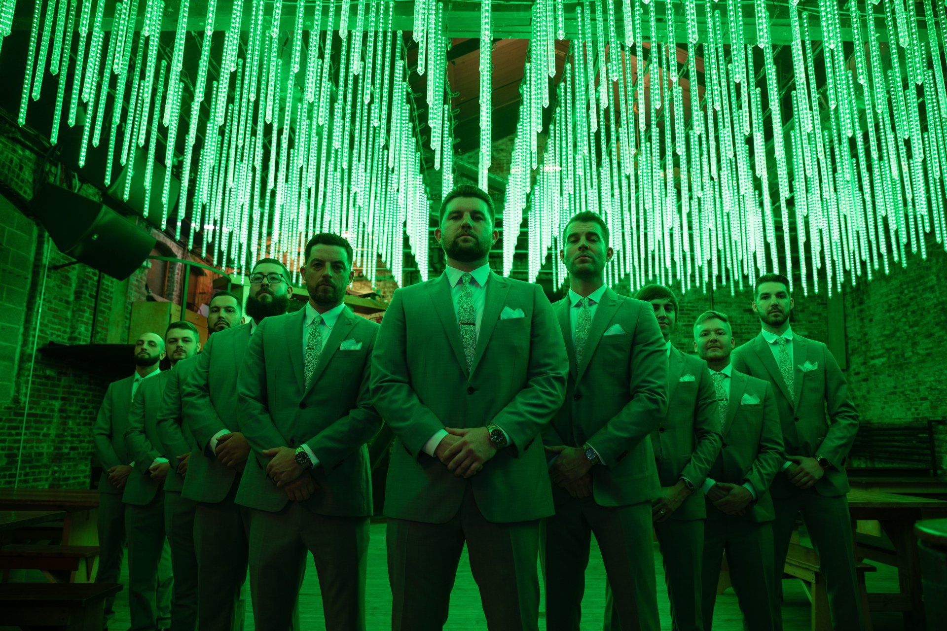 Group of men in suits standing beneath green neon lights in a dramatic indoor setting