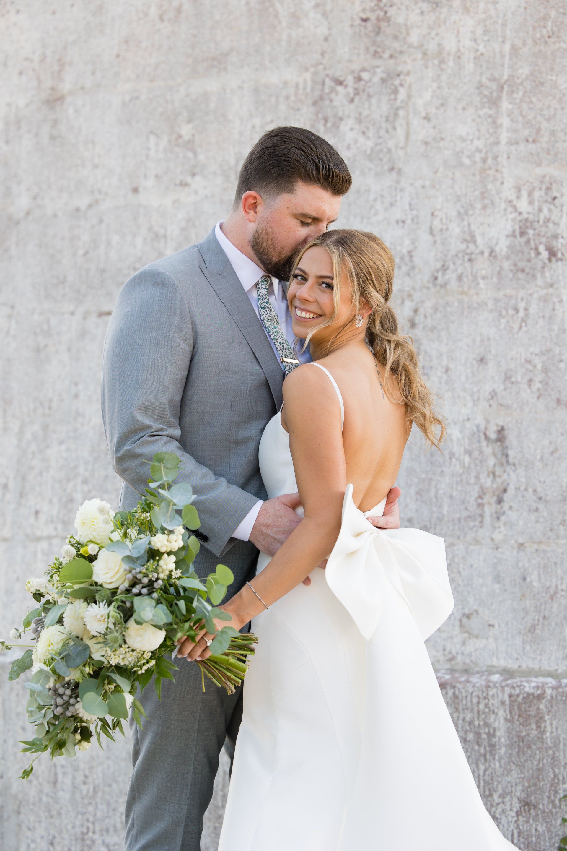 Wedding couple posing against a stone wall, groom in gray suit and bride in white dress holding a bouquet