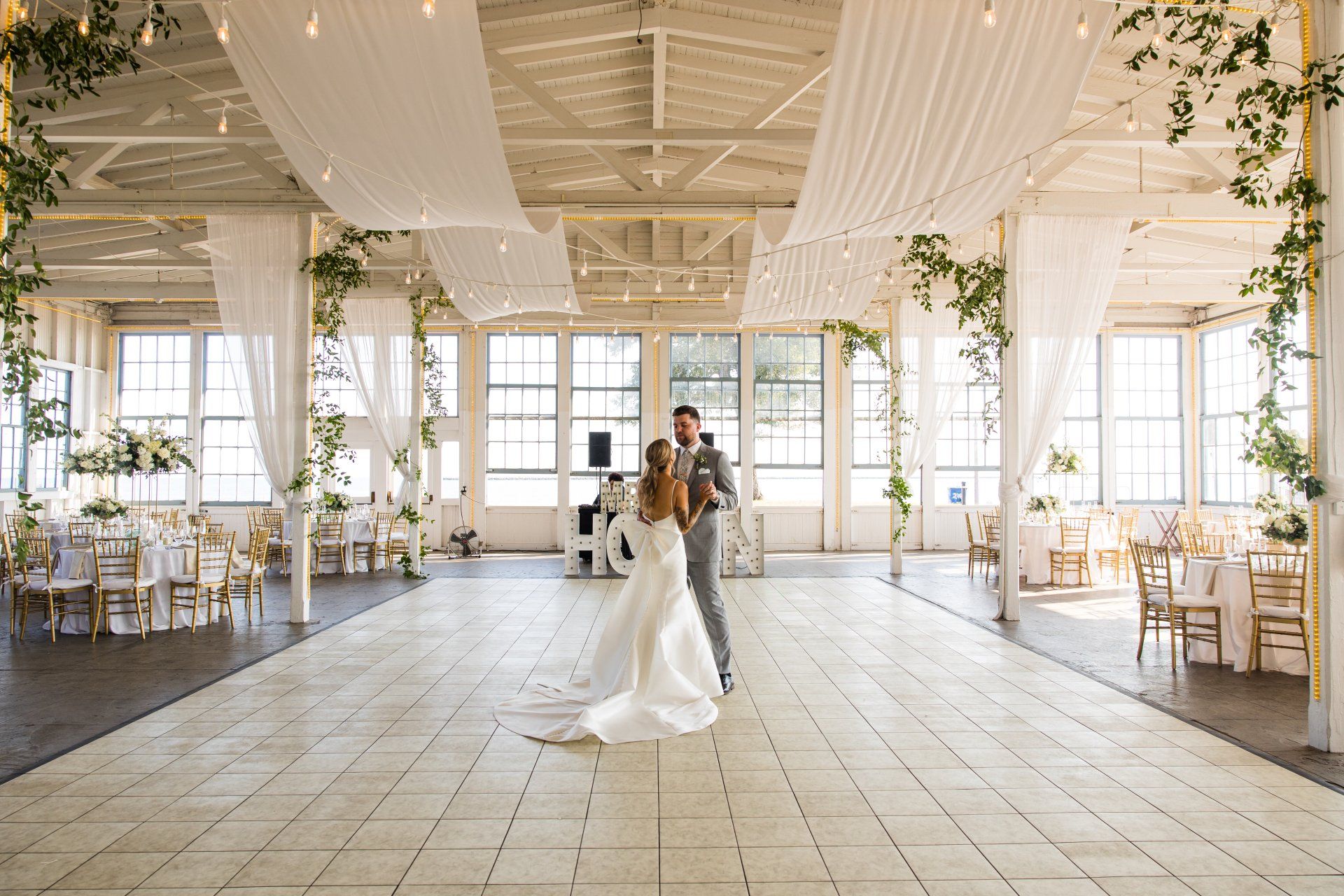 Bride and groom dancing in a bright white wedding hall with draped fabric and chandeliers.