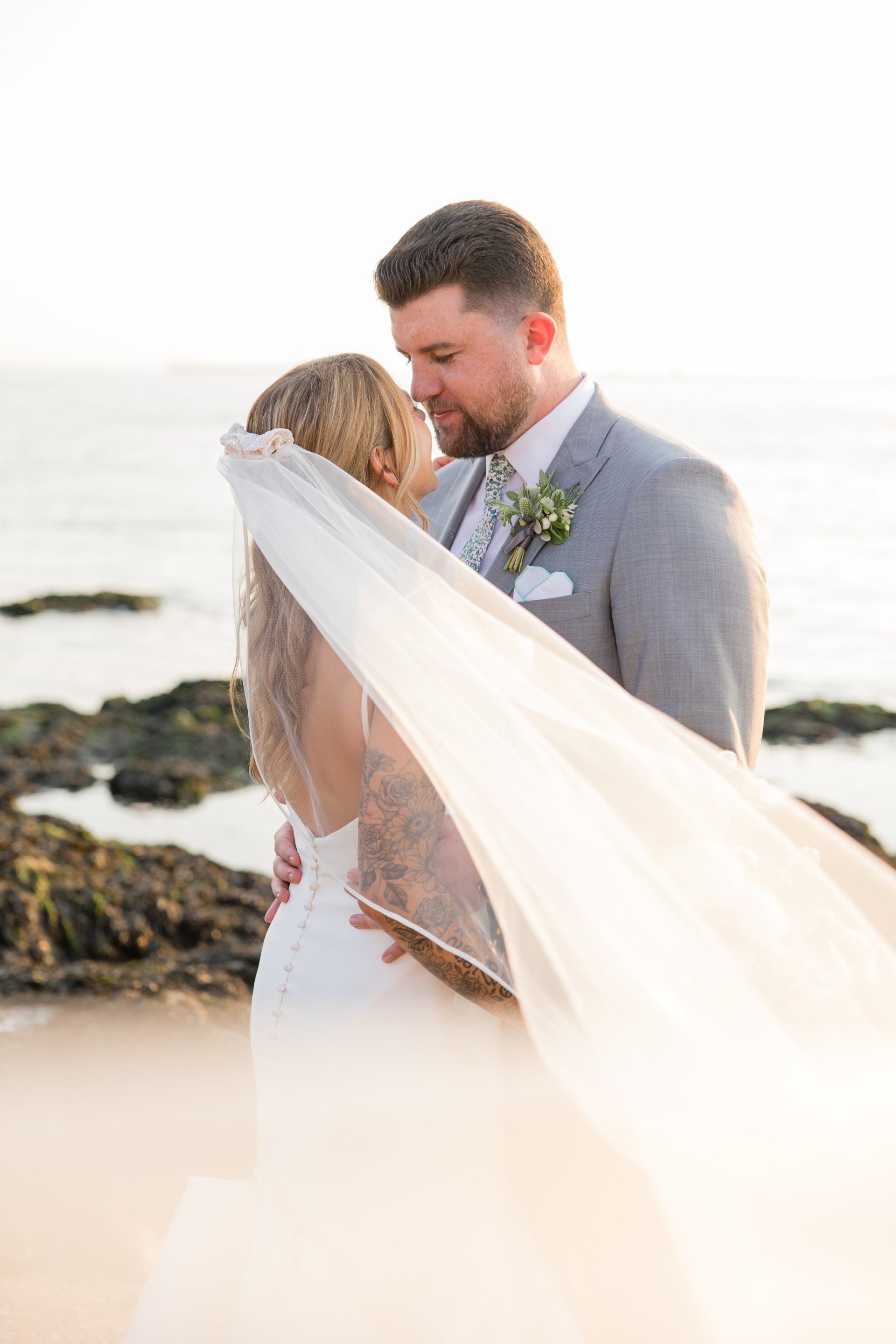 Bride and groom embracing on a beach, with the bride’s veil flowing in the wind