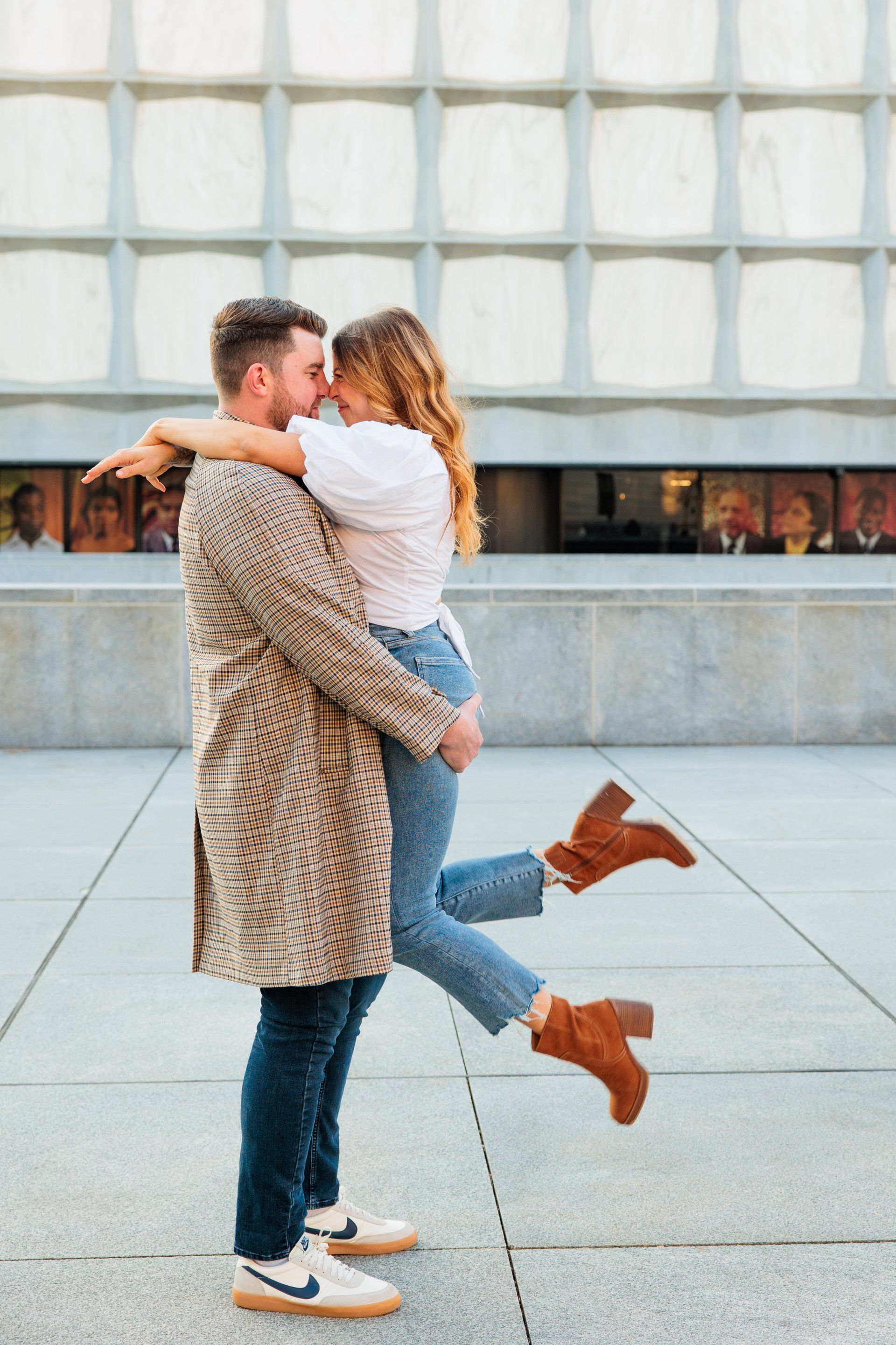 Couple embracing outdoors, one lifting the other in front of a modern building.