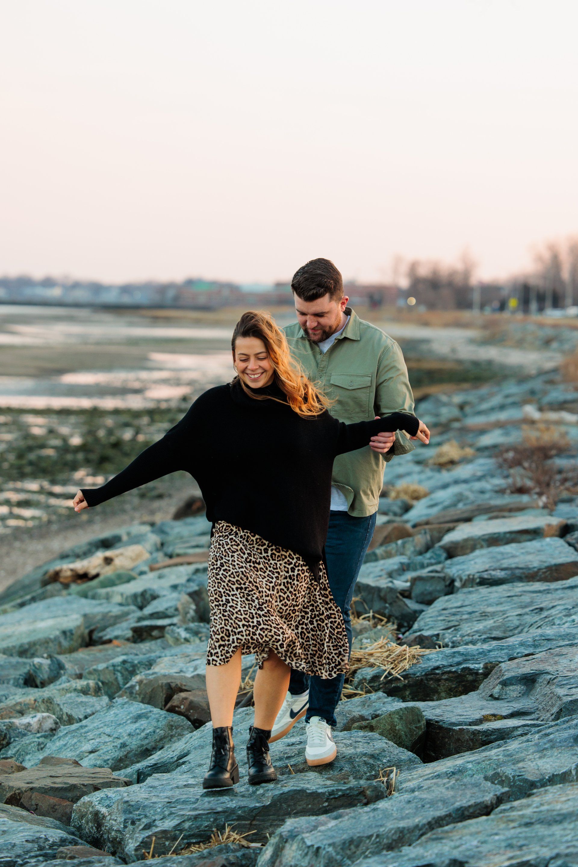 Couple walking hand-in-hand on rocky shoreline at sunset, smiling and balancing together