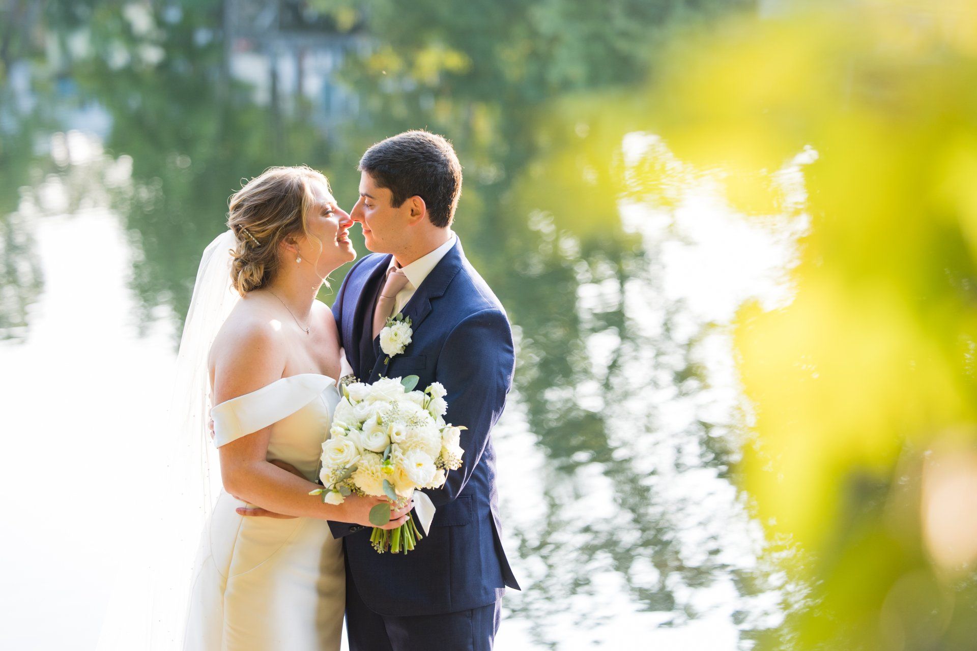 Bride and groom embracing outdoors by a bright lake, holding a white bouquet.