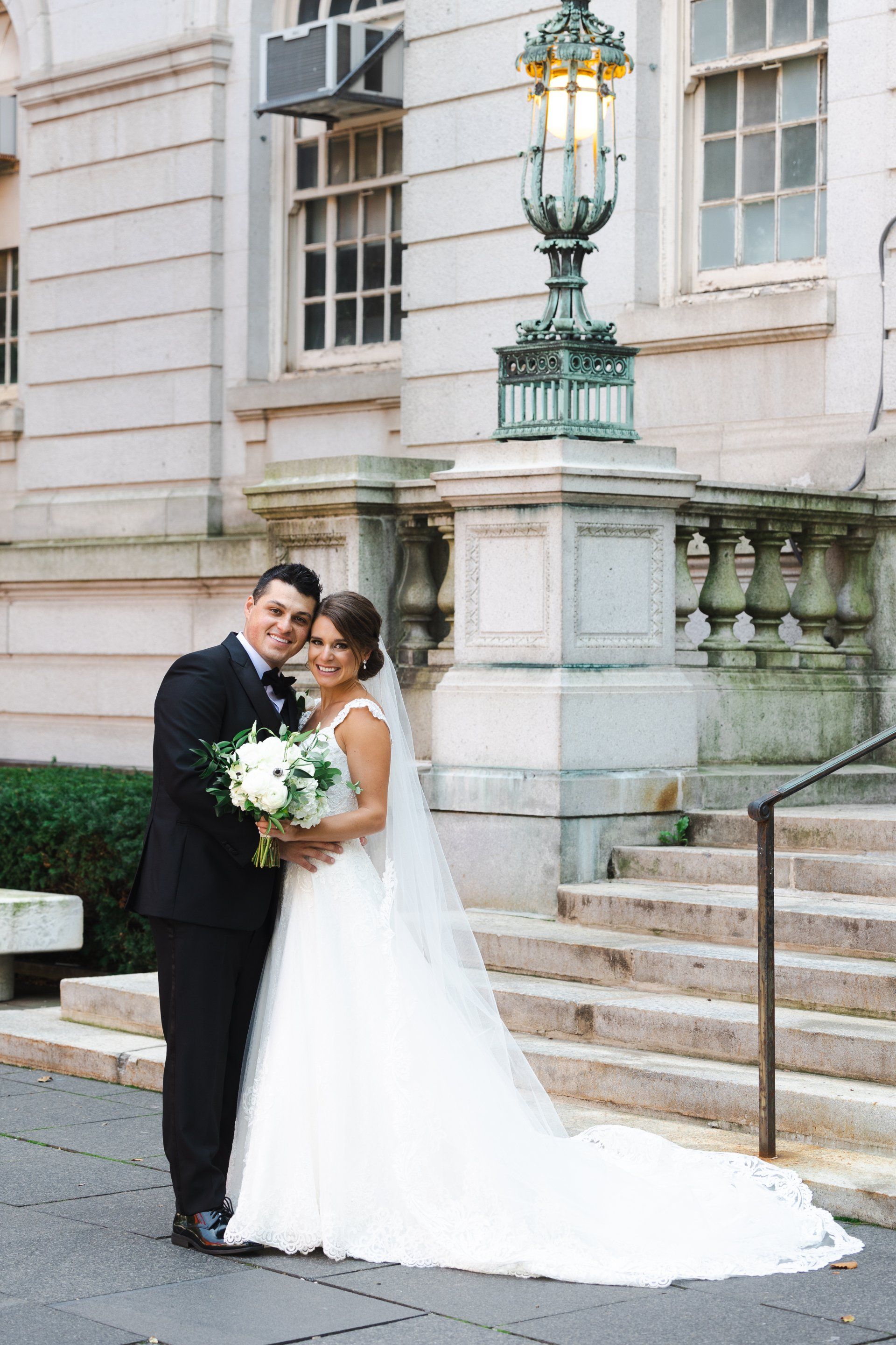 Wedding couple posing outside a stone building, bride in white gown holding bouquet, groom in black tuxedo.