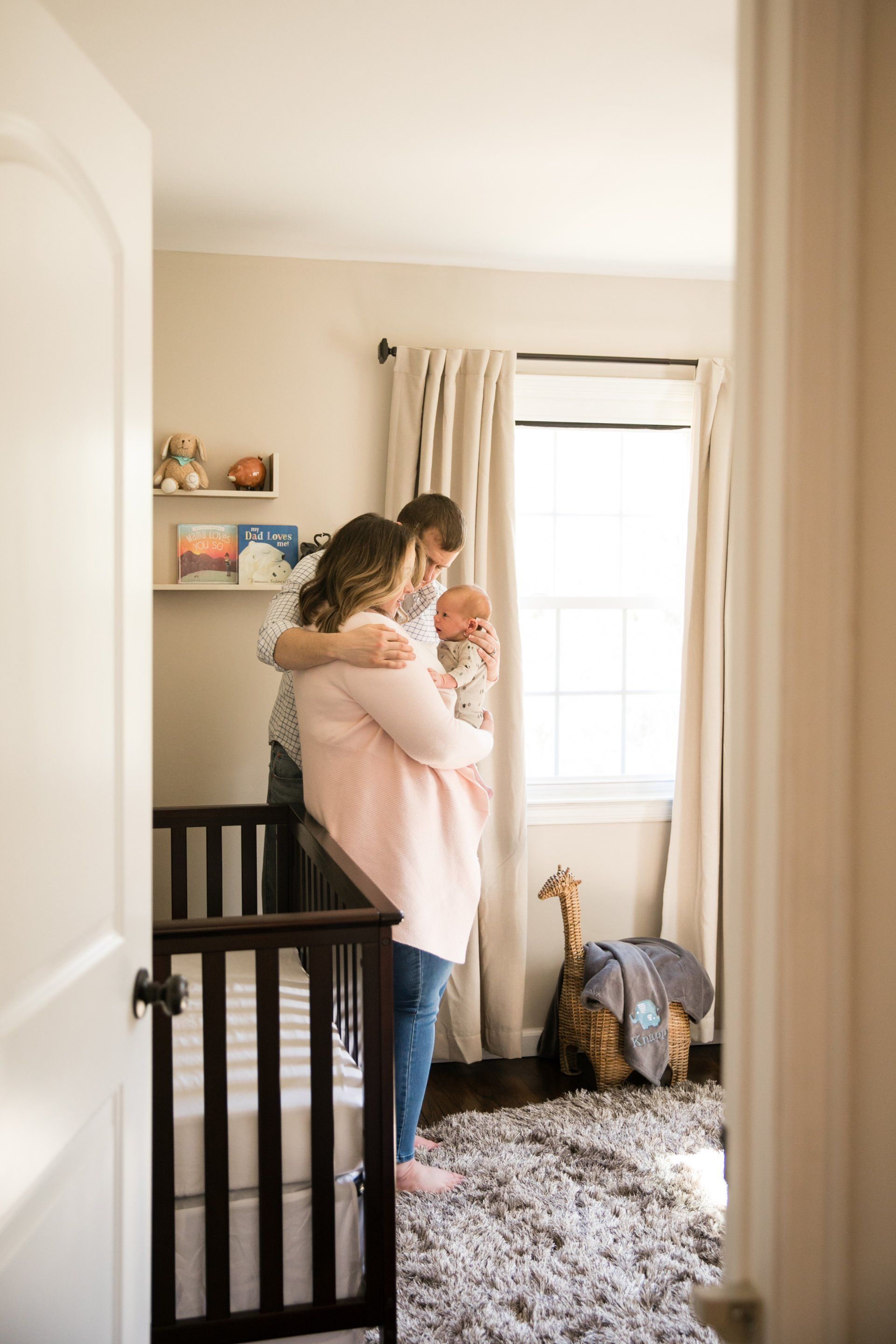Adult holding a toddler in a nursery by a bright window, with a crib and stuffed backpack nearby.