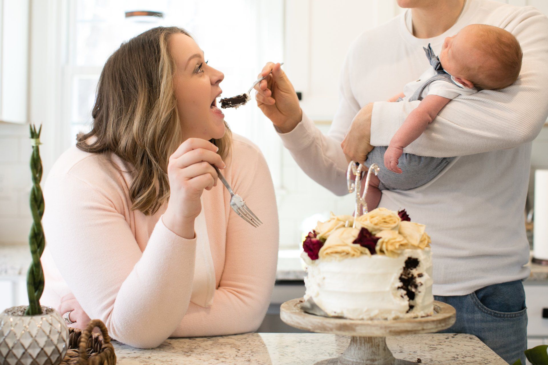 Woman feeds frosting to a smiling child beside a cake while another adult holds a baby in a kitchen.