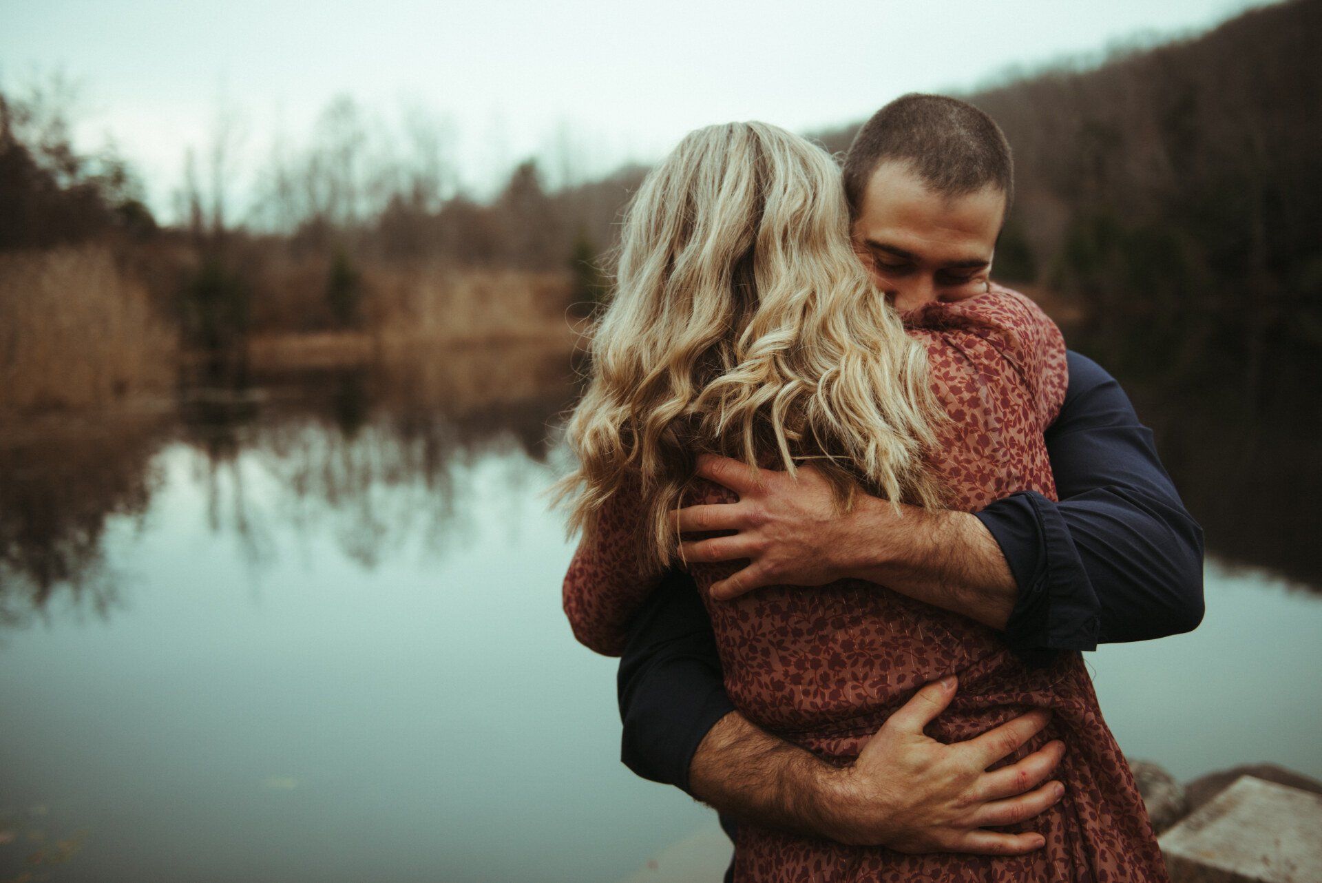 Couple embracing by a calm lake, with bare trees and a soft, muted background.