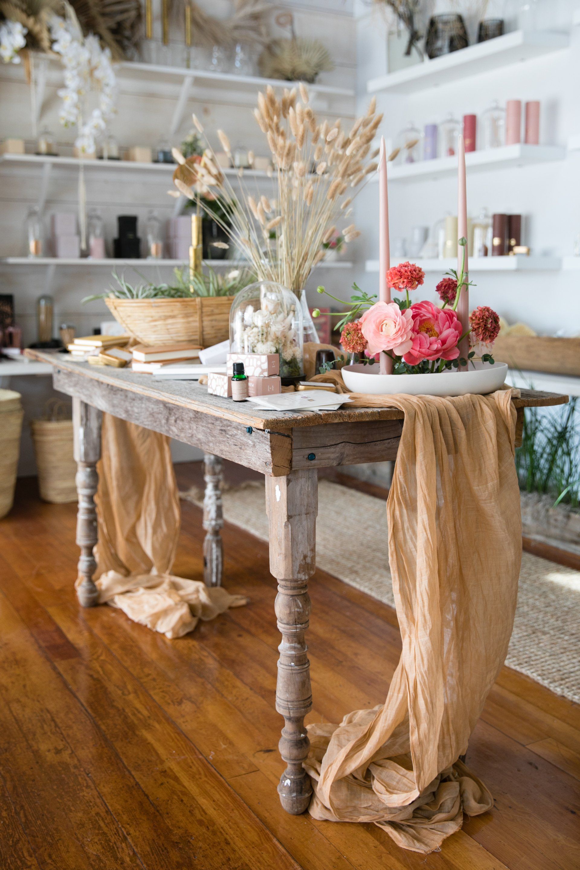 Rustic wooden table with flowers, dried grasses, and beige draped fabric in a bright room