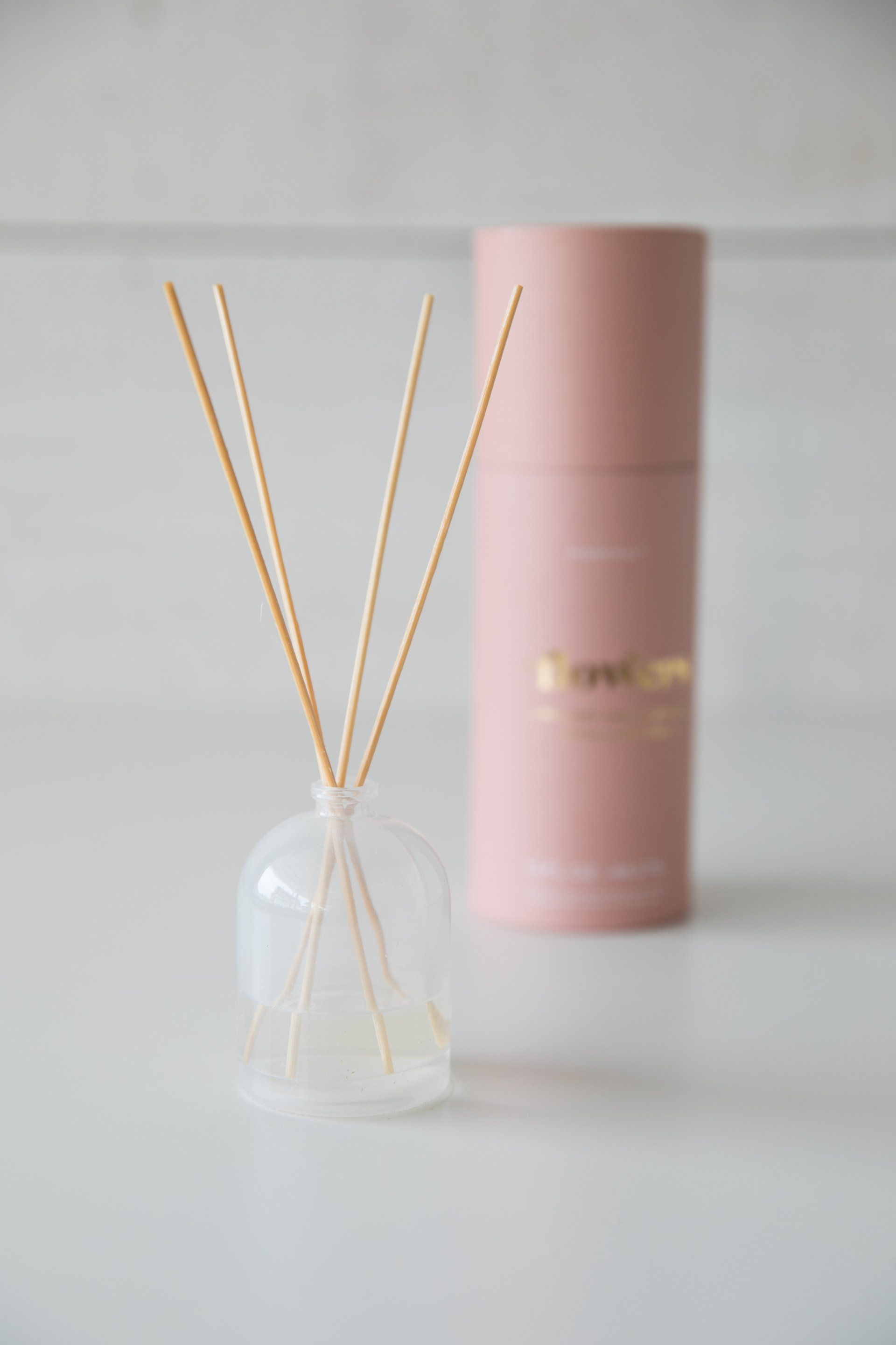 A pink cosmetic tube beside a white reed diffuser with golden sticks on a light background