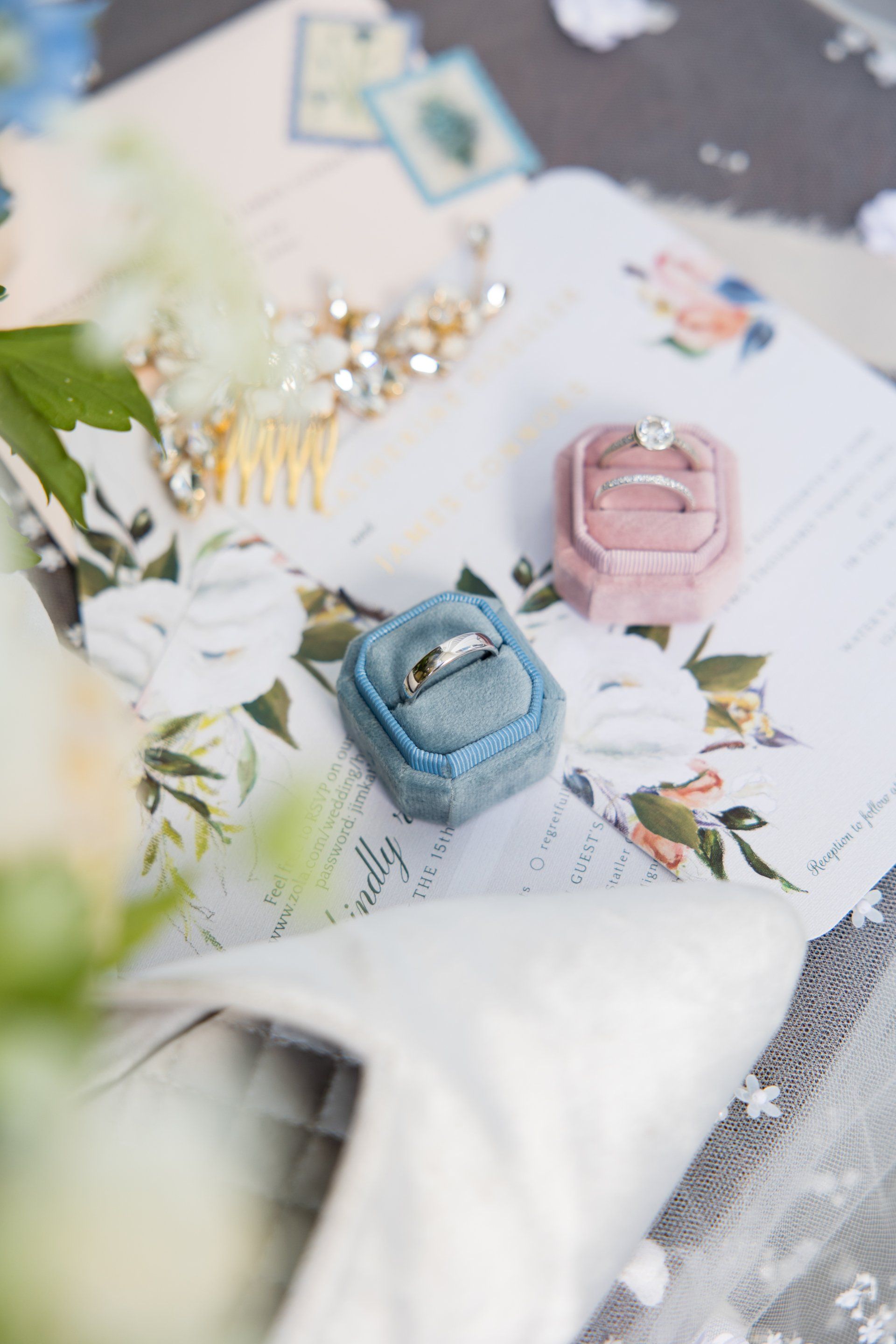 Two square gemstone rings, one blue and one pink, on a floral surface with a white flower and jewelry.