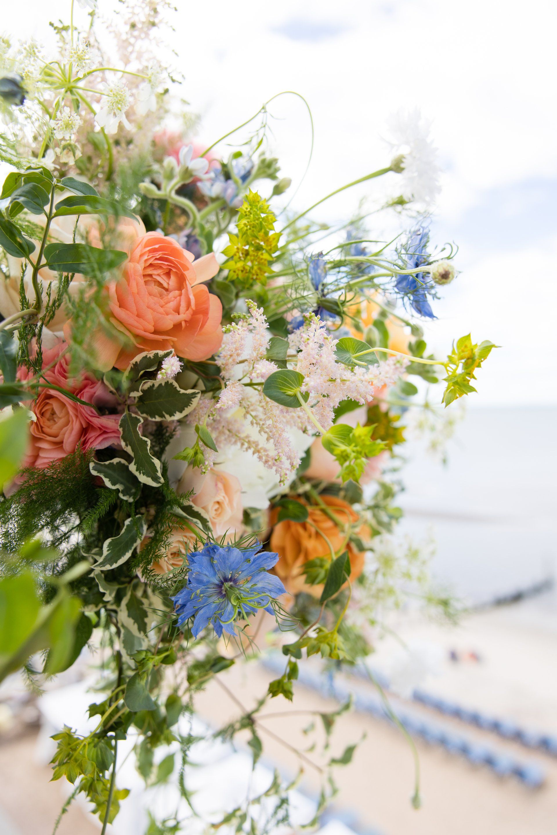 Pastel wedding bouquet with peach, blue, and white flowers against a bright seaside backdrop