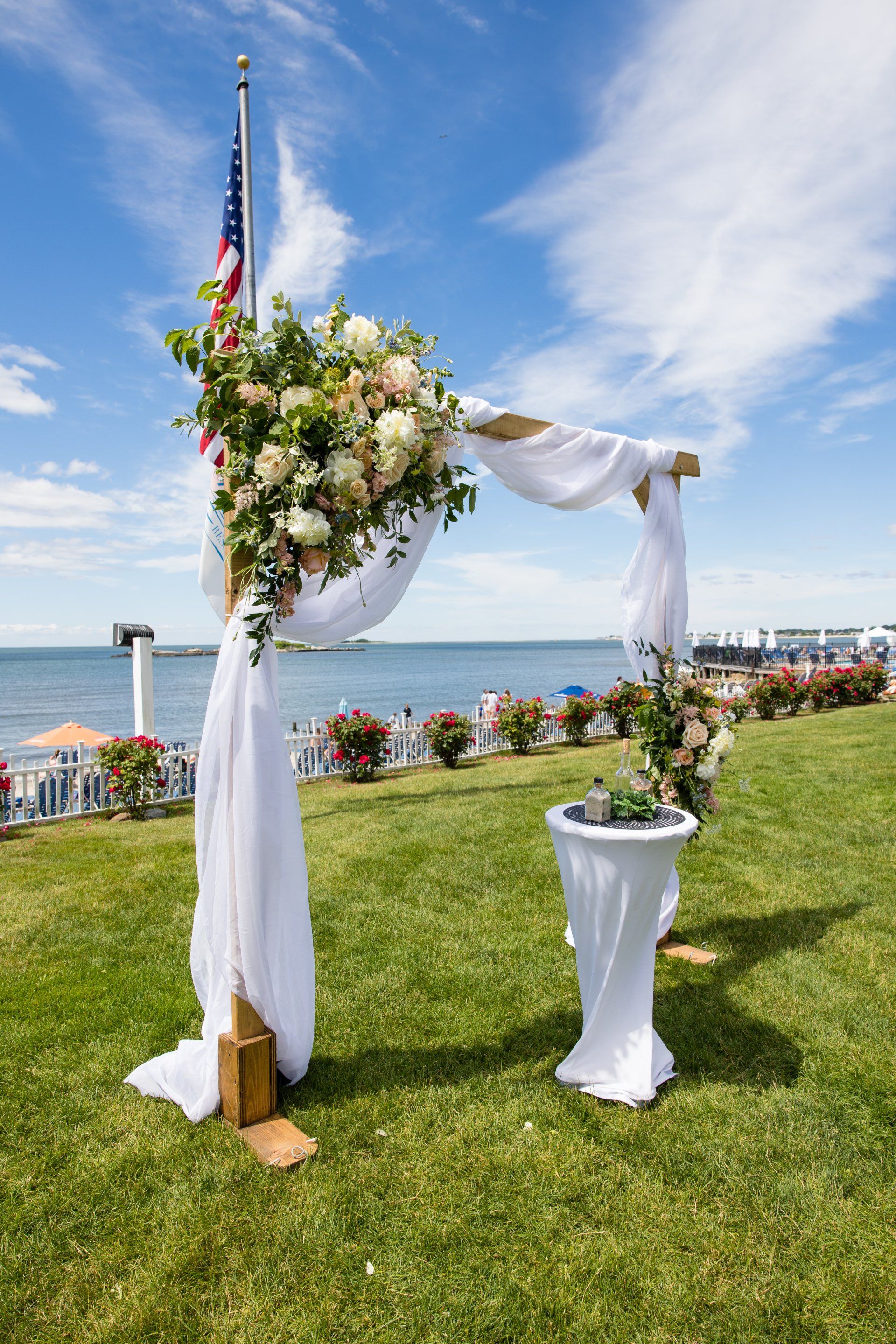 White floral wedding arch and table on a grassy waterfront lawn under a blue sky