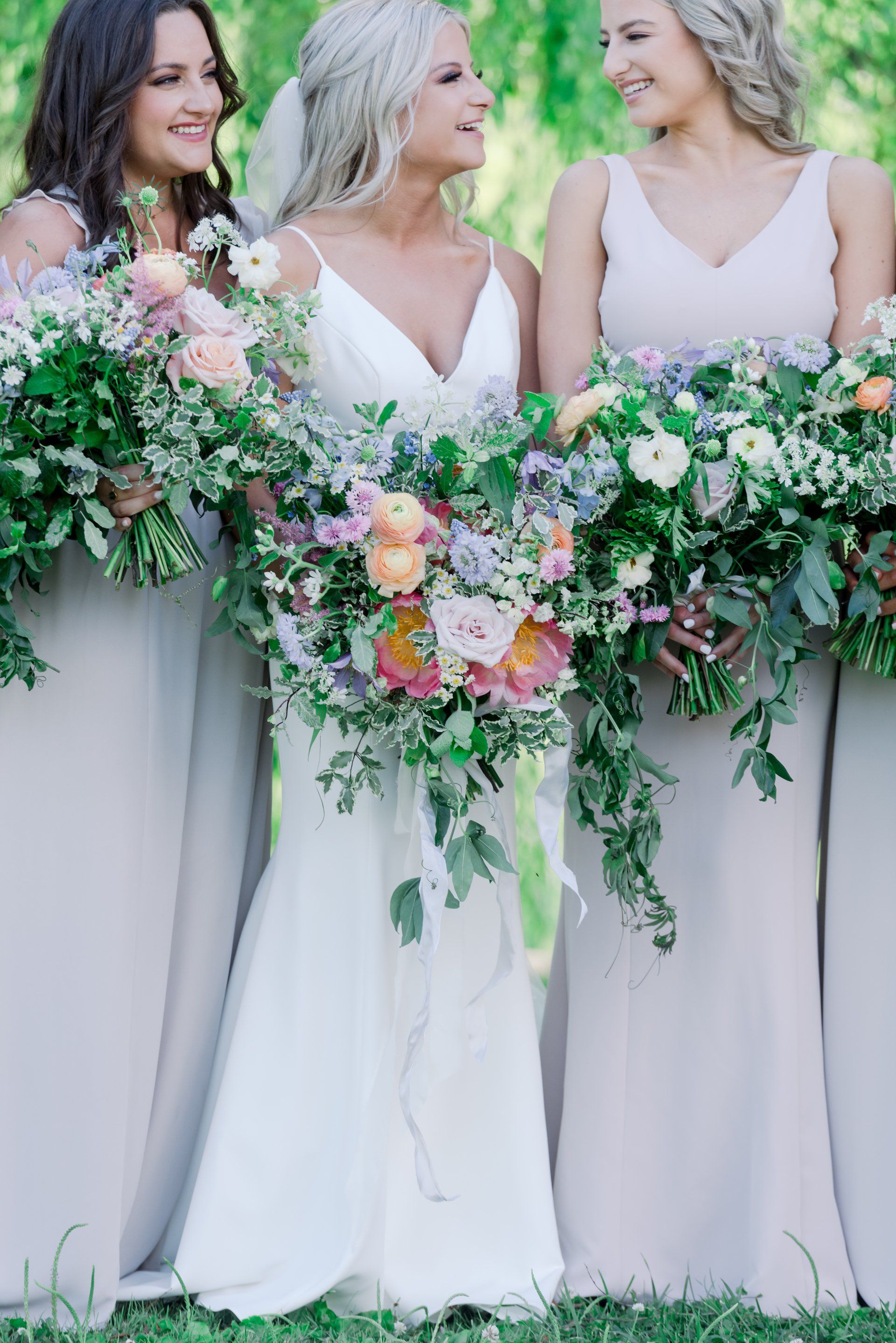 Three brides in white dresses holding lush pink and green bouquets outdoors