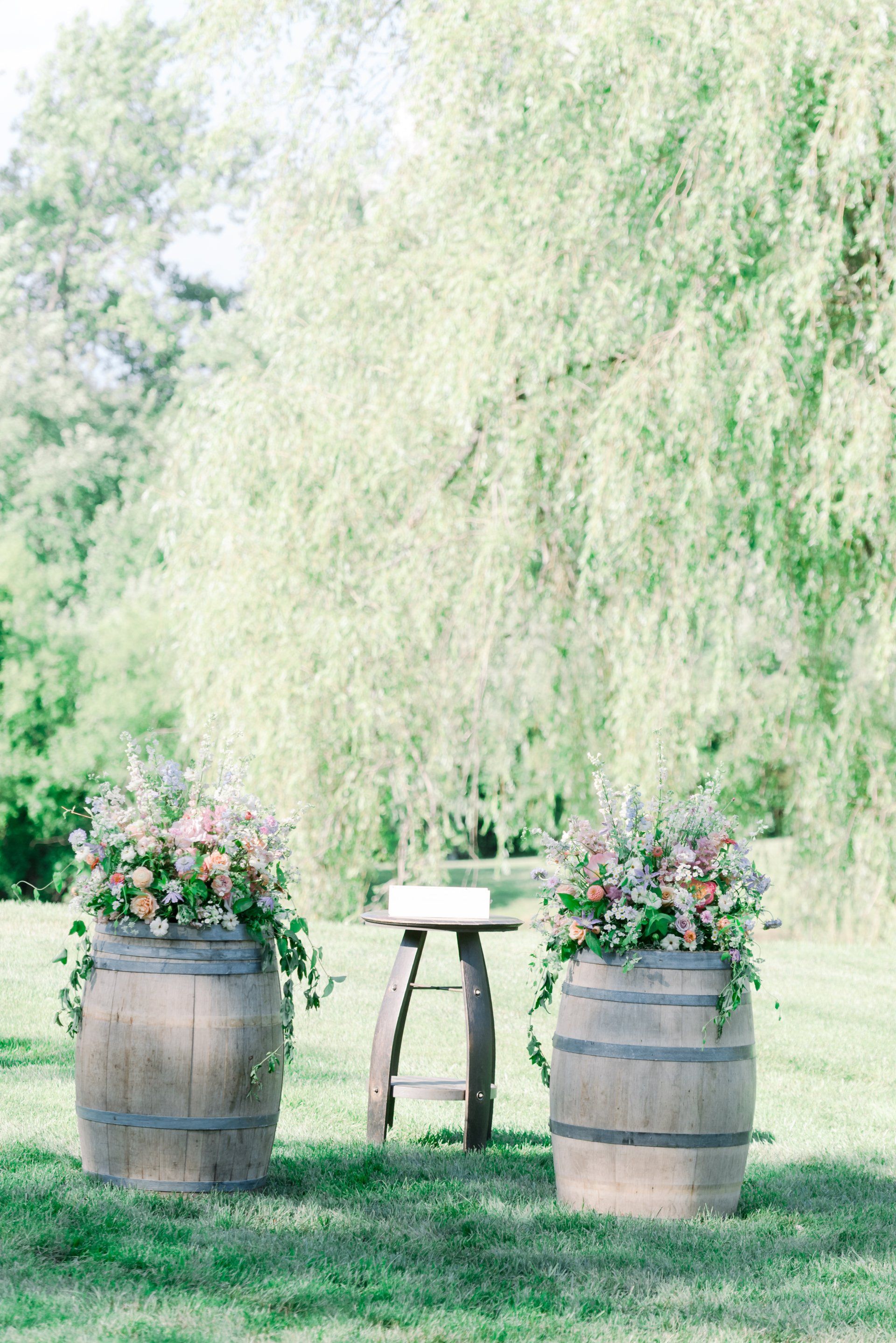 Rustic garden setup with two flower-filled barrels and a small table under a leafy tree.