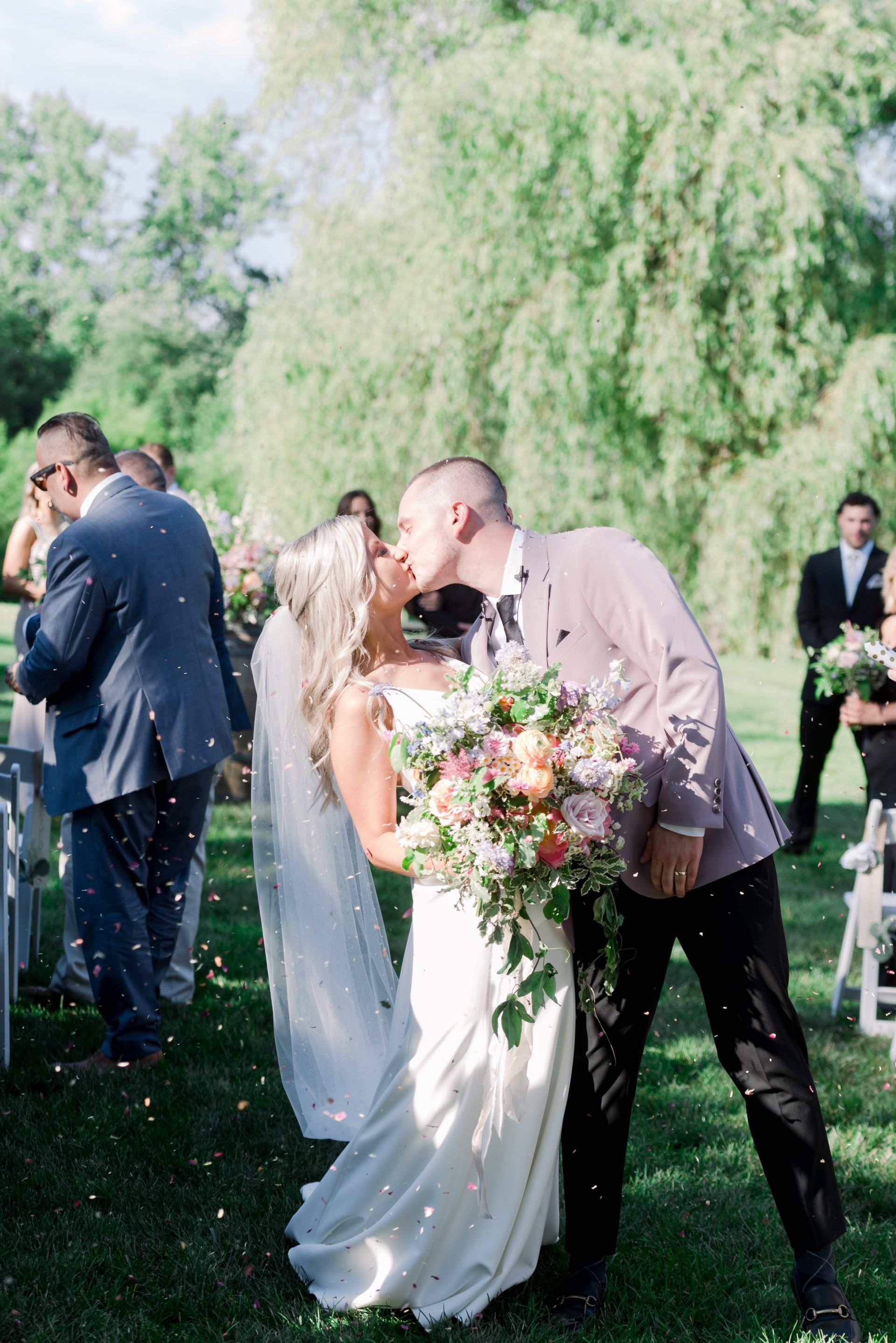 Newlyweds kissing outdoors in a sunlit garden, bride holding a bouquet and wearing a white dress