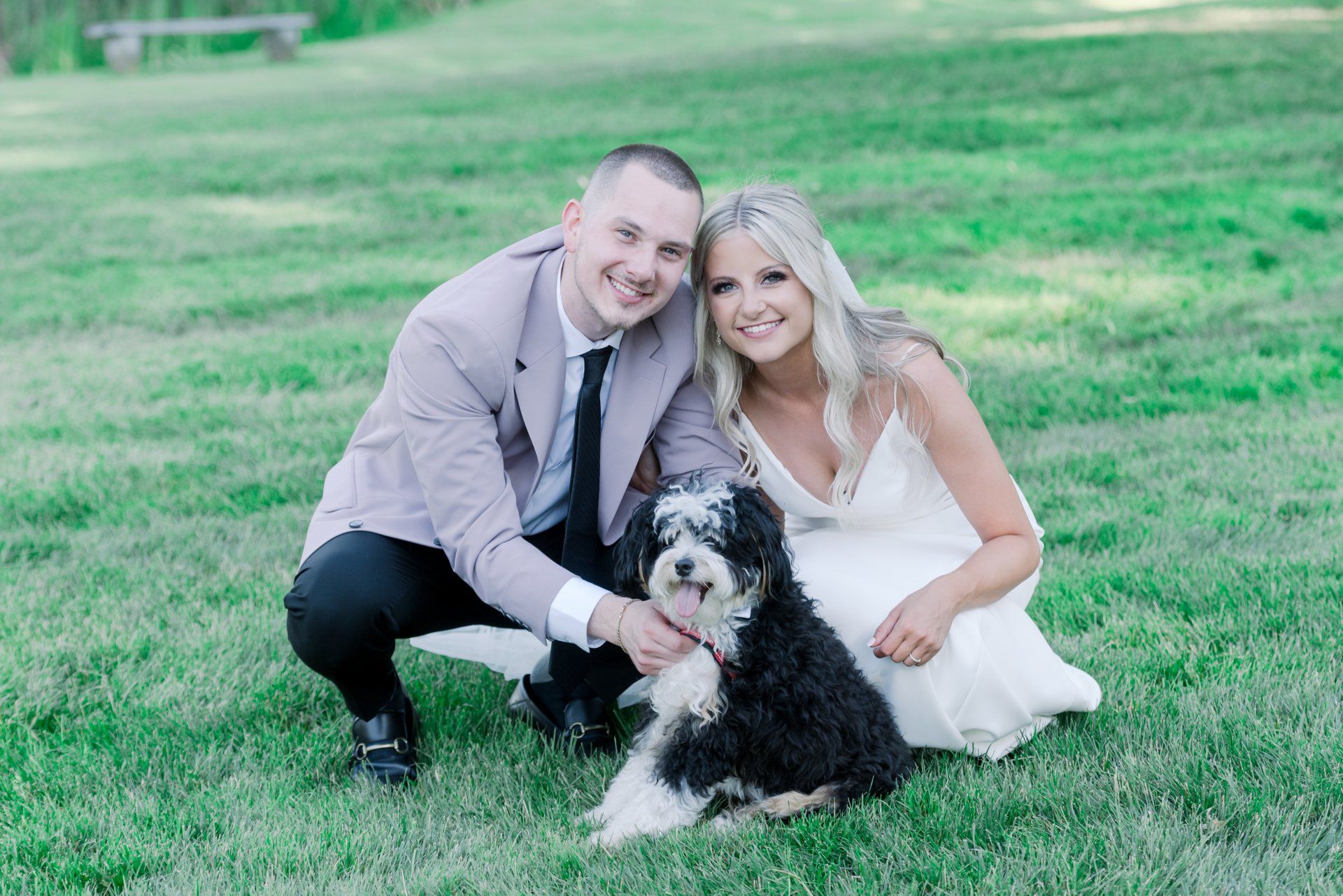 Couple in formal clothes kneeling on grass with a black-and-white dog, smiling outdoors