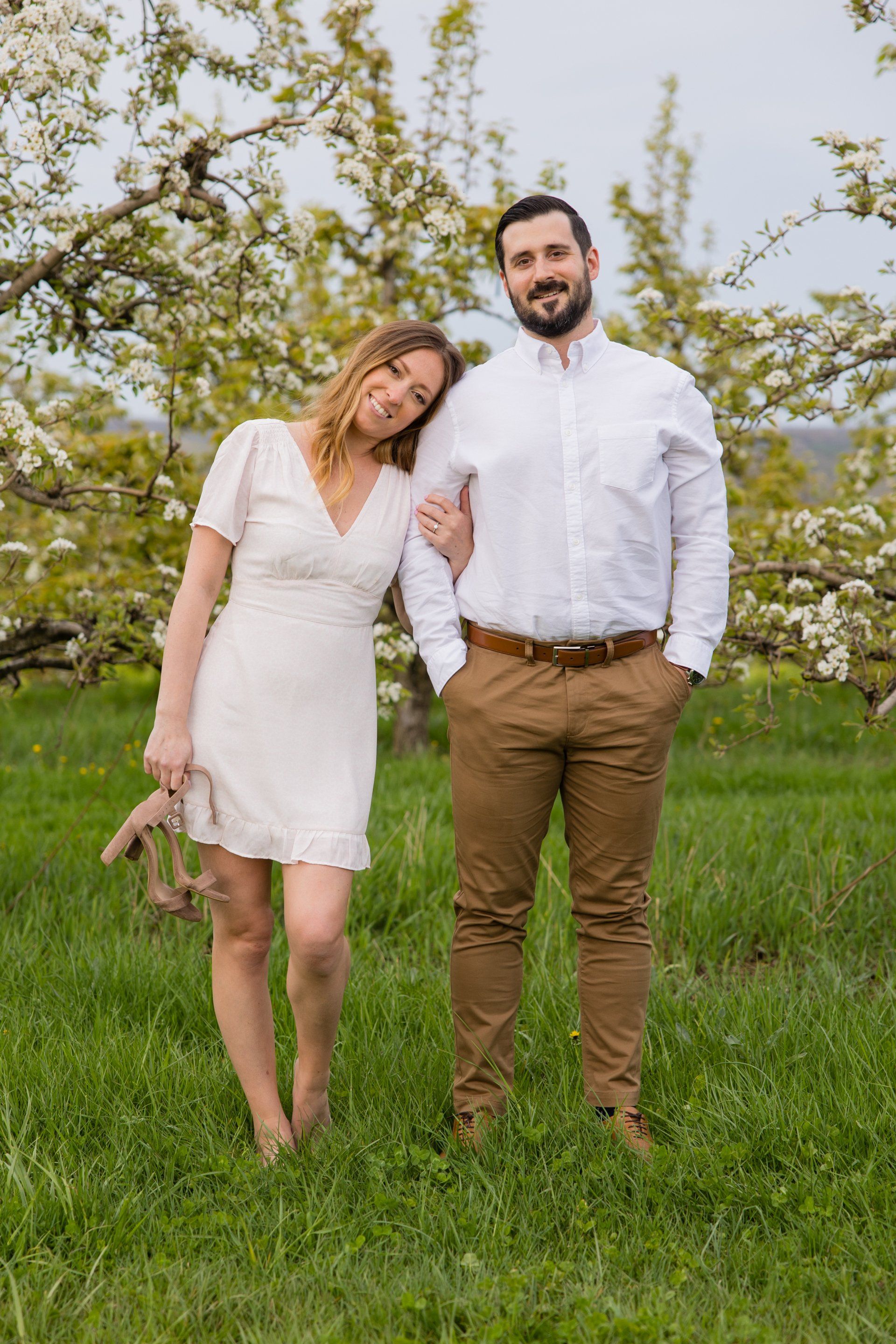 Two people standing in a grassy orchard, smiling, with blooming trees behind them.