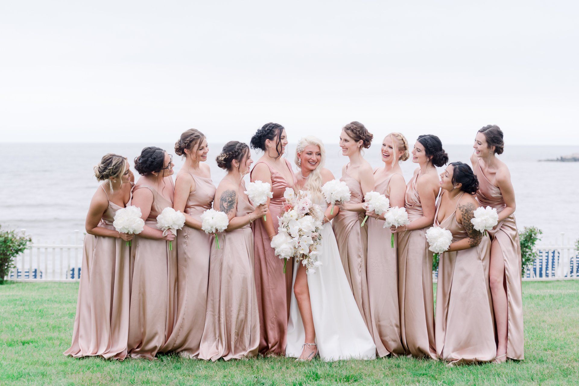 Bridal party posing outdoors by the water, in blush and white dresses with bouquets.