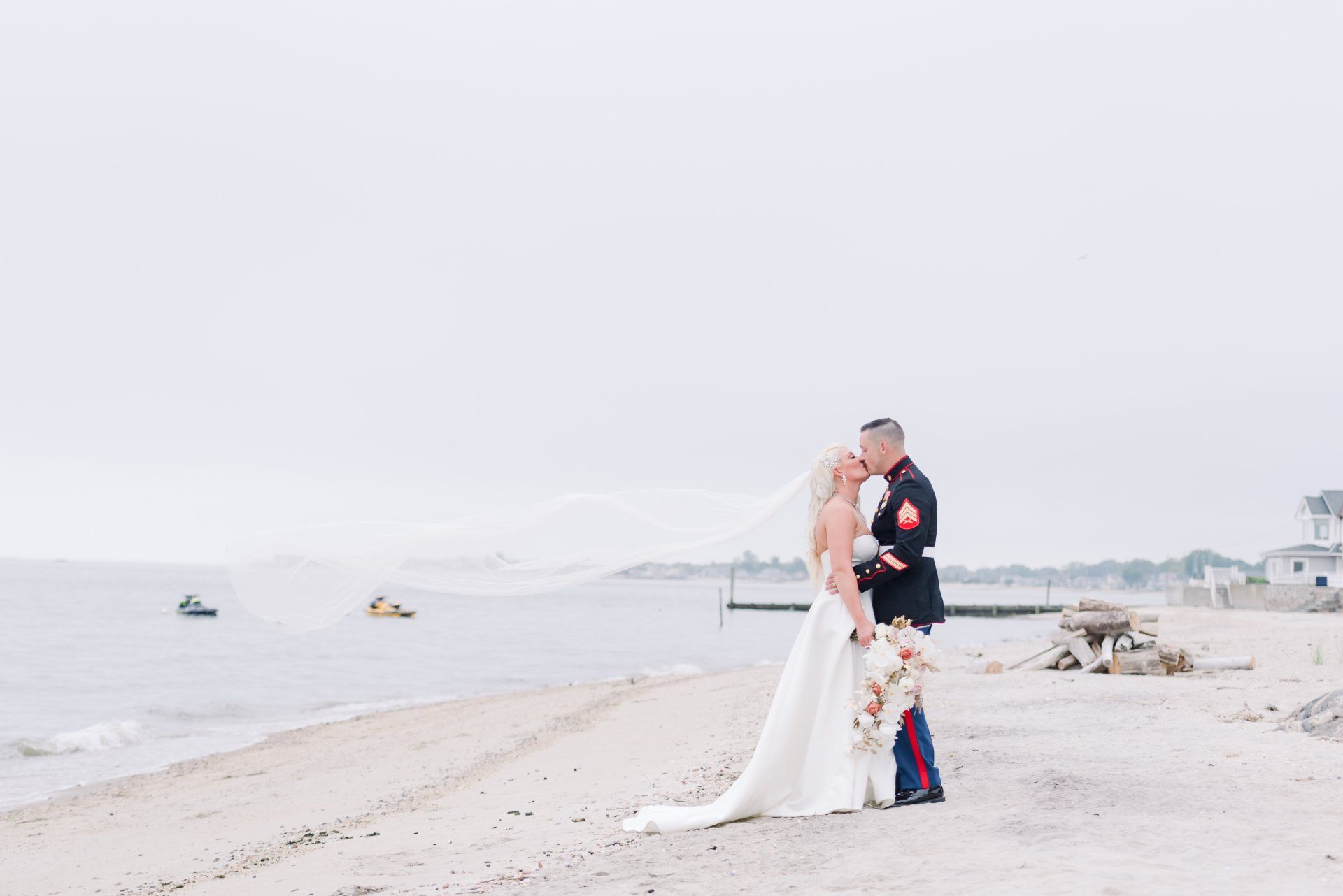 Bride and groom embracing on a bright beach beside the sea, with boats in the background