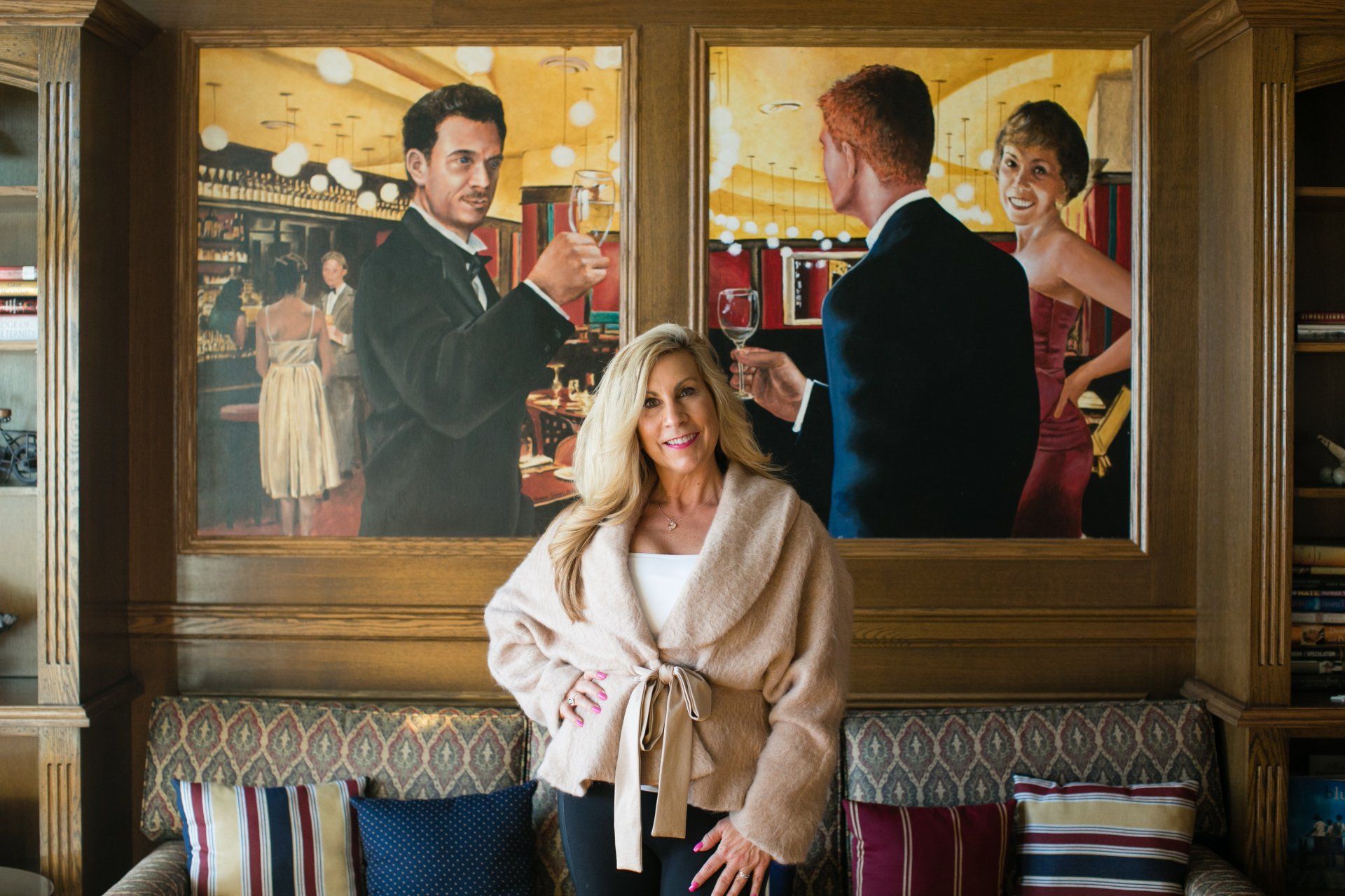 Woman in a cream robe sits before a large painting of three people holding drinks.