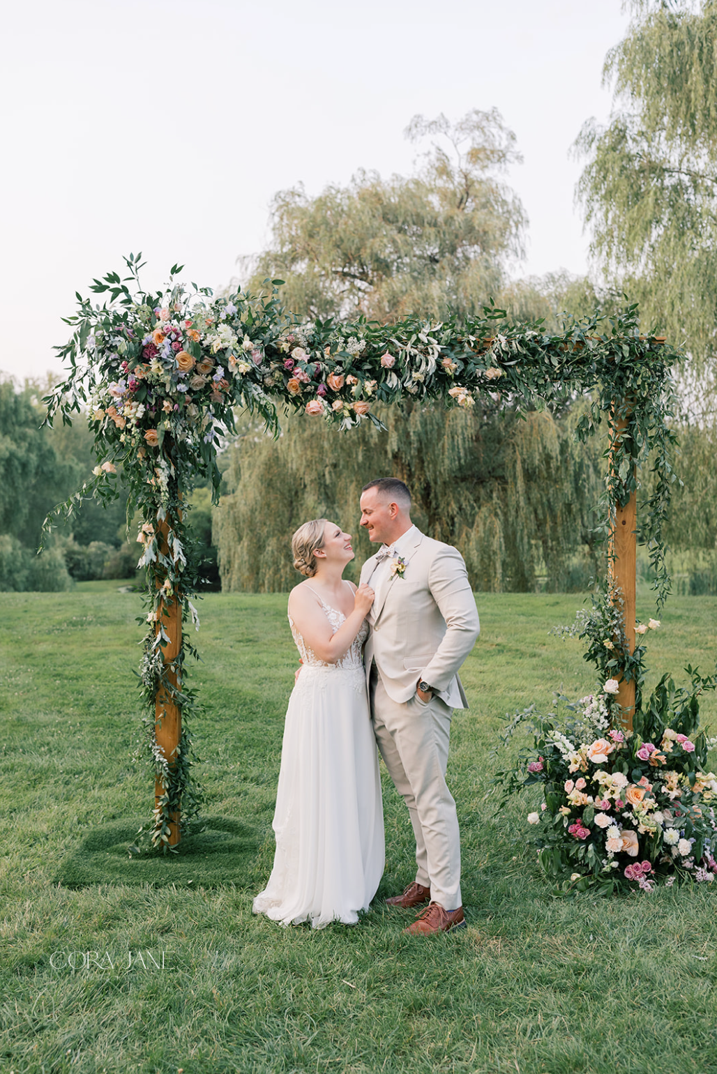 Couple in formal attire under a floral wedding arch in a green garden, sharing an intimate moment